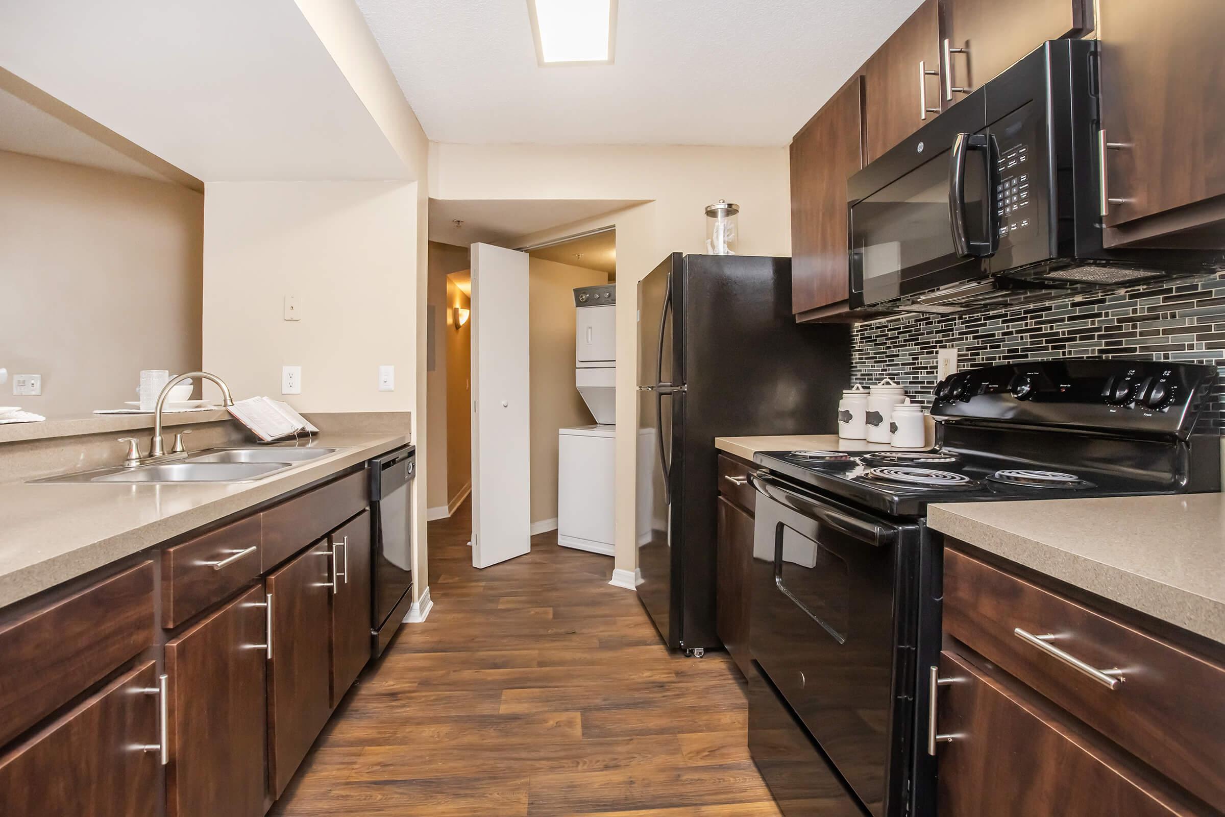 Modern kitchen featuring dark wood cabinetry, stainless steel appliances including a microwave and stove, and a contrasting countertop. The space has a laminate floor and a tile backsplash, with an open layout leading to an adjacent room. Natural light comes from a ceiling fixture.
