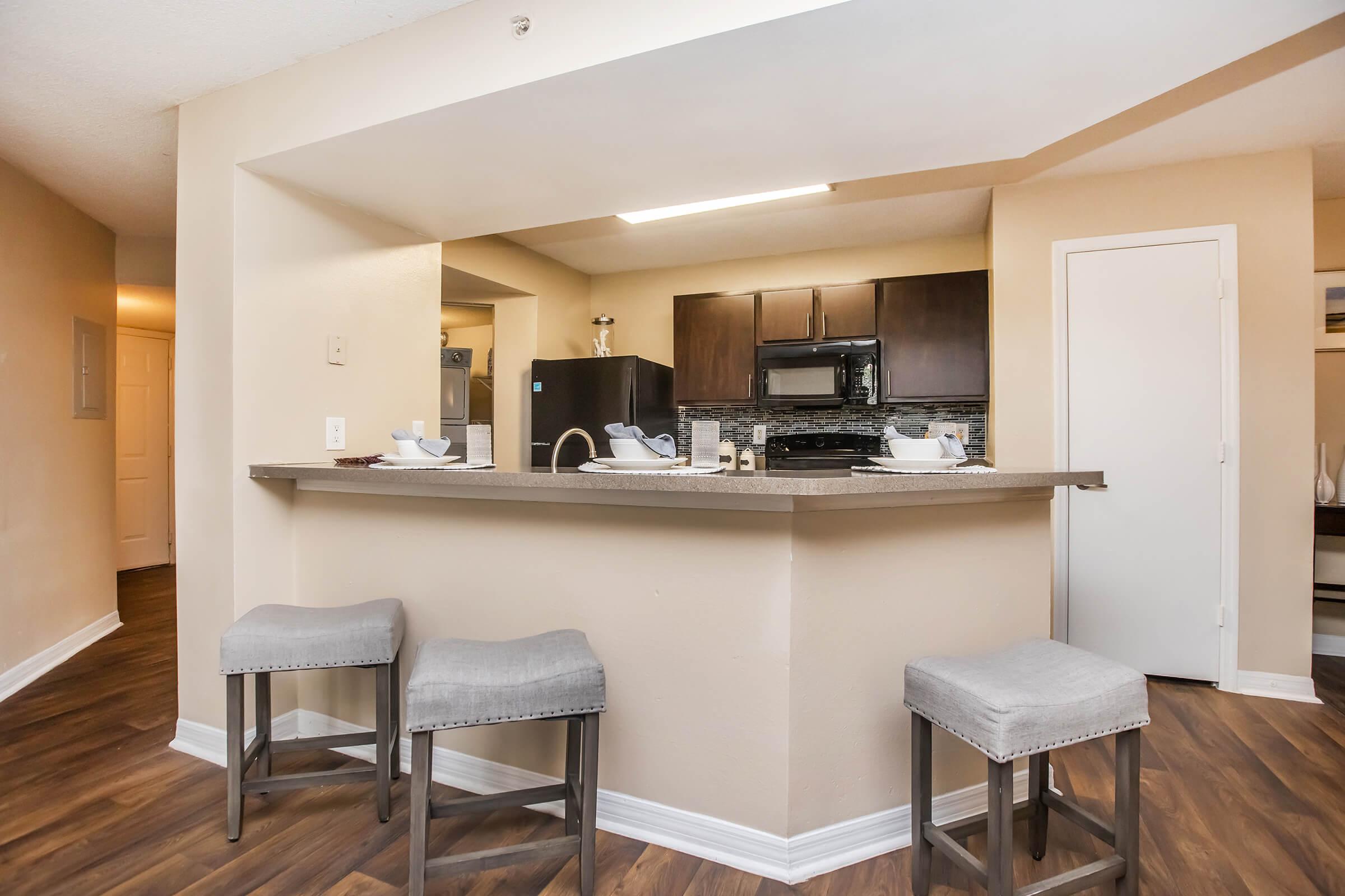 Modern kitchen interior featuring a bar area with three stools, a granite countertop, and dark wood cabinetry. The kitchen is equipped with a black microwave and oven, and there is neutral-colored wall paint. The floor has a wood-like finish, creating a warm and inviting atmosphere.