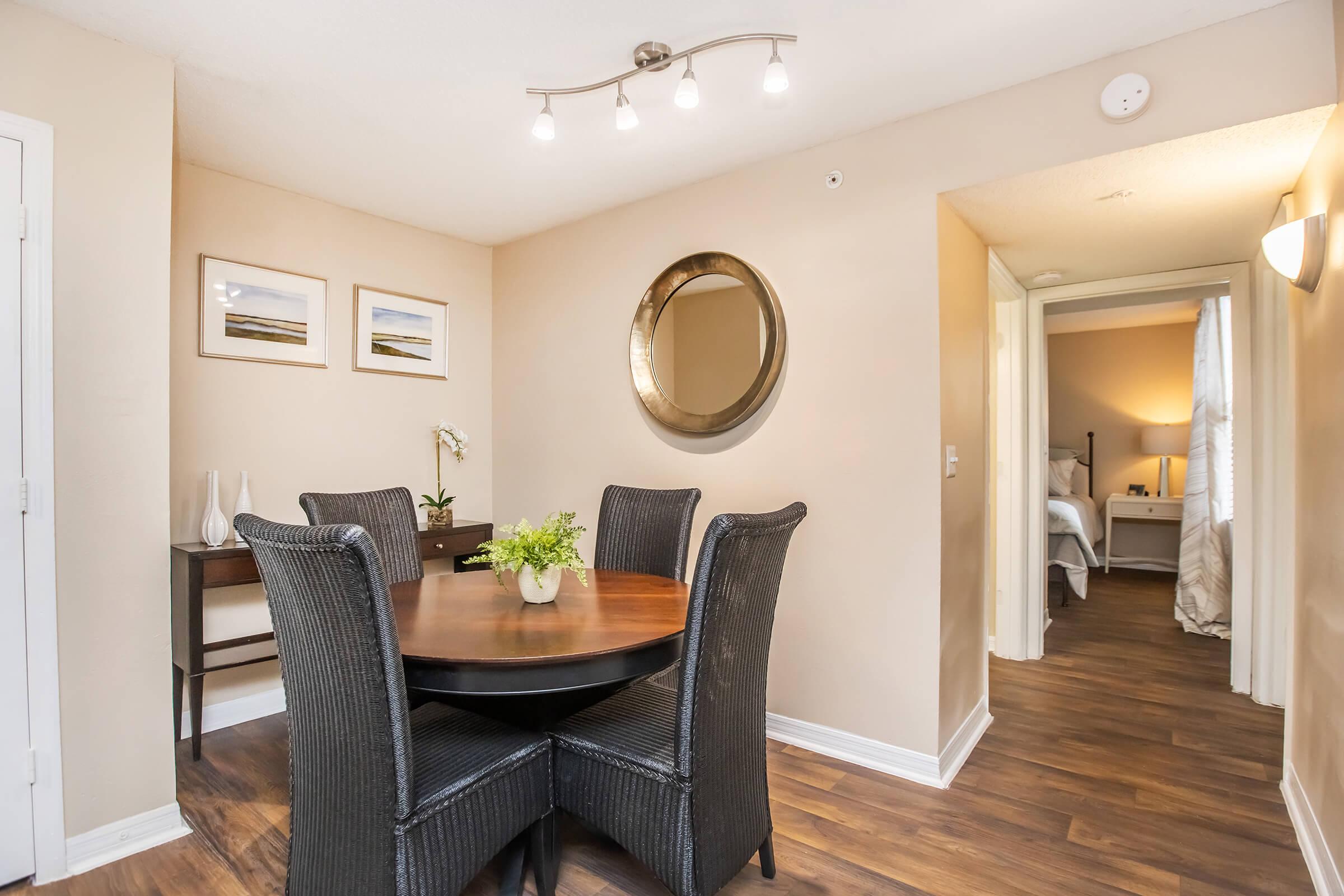 A cozy dining area featuring a round wooden table with four black wicker chairs, a small side table, and a decorative vase with a plant. The walls are painted in a warm beige tone, and there are framed pictures and a round mirror. A doorway leads to a softly lit bedroom in the background.