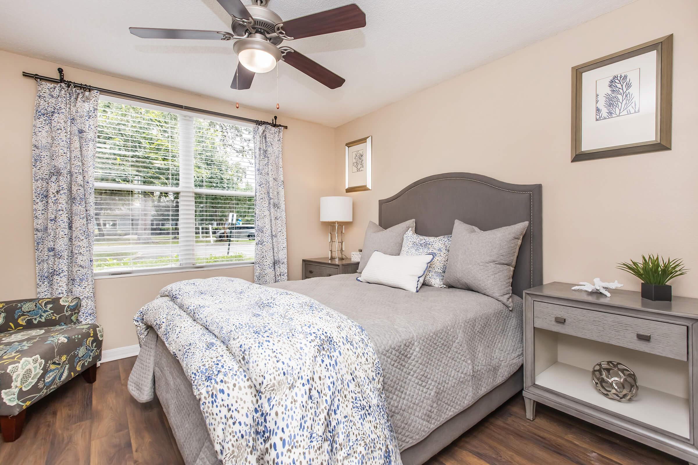 A cozy bedroom featuring a queen-sized bed with gray bedding and decorative pillows, a nightstand with a lamp, and a small decorative plant. The room has natural light coming through a window with floral curtains and a comfortable accent chair in the corner. The walls are a soft beige color.