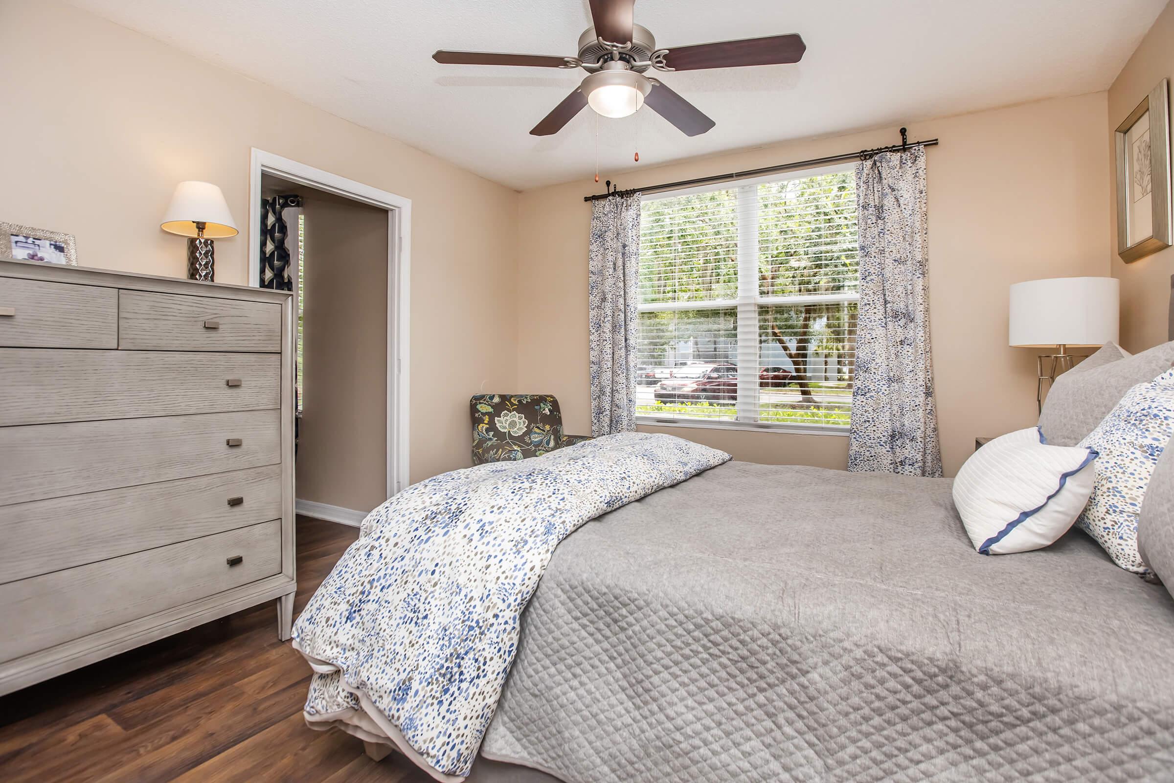 Cozy bedroom featuring a queen-sized bed with a patterned throw blanket, a dresser, and a chair near the window. Natural light streams in through the window, and soft blue and white decor accents create a serene atmosphere. A ceiling fan adds to the comfort of the space.