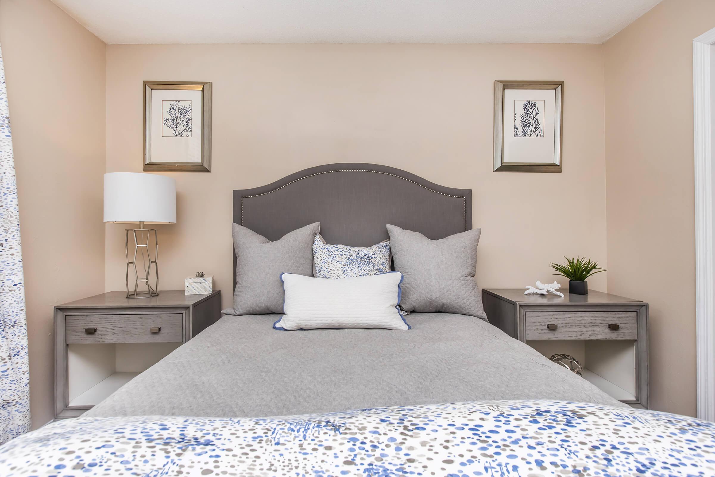 A cozy bedroom featuring a gray upholstered headboard and neutral bedding. The walls are painted a soft beige, and there are two decorative framed prints above the bed. Each side of the bed has a gray nightstand with a small lamp and a potted plant. The overall decor is modern and inviting.