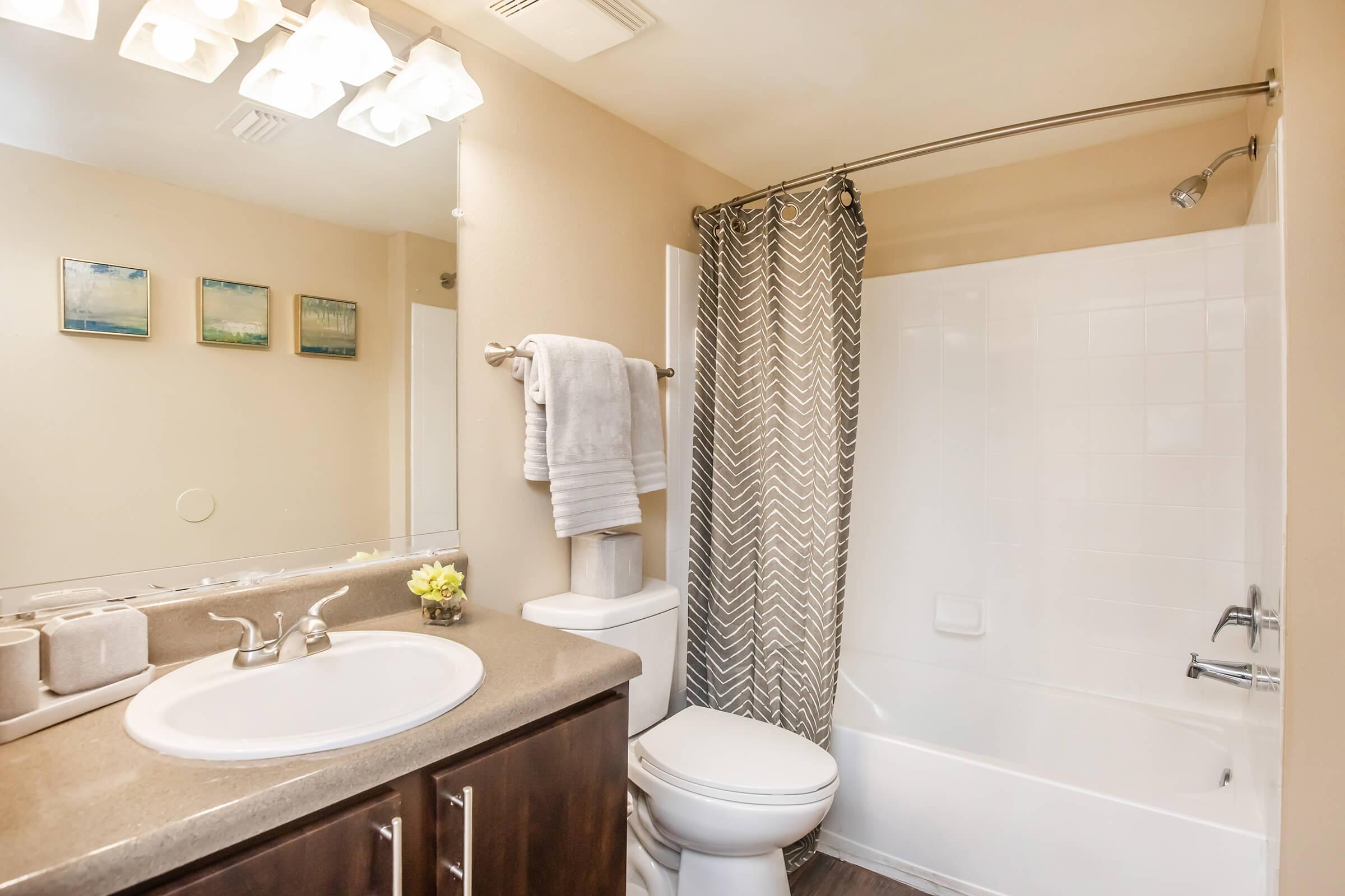 A clean and modern bathroom featuring a white bathtub with a shower curtain, a round sink with a countertop, a toilet, and a large vanity mirror. The walls are painted in a light color, and there are three small framed pictures above the sink. A vase with flowers adds a touch of decor.