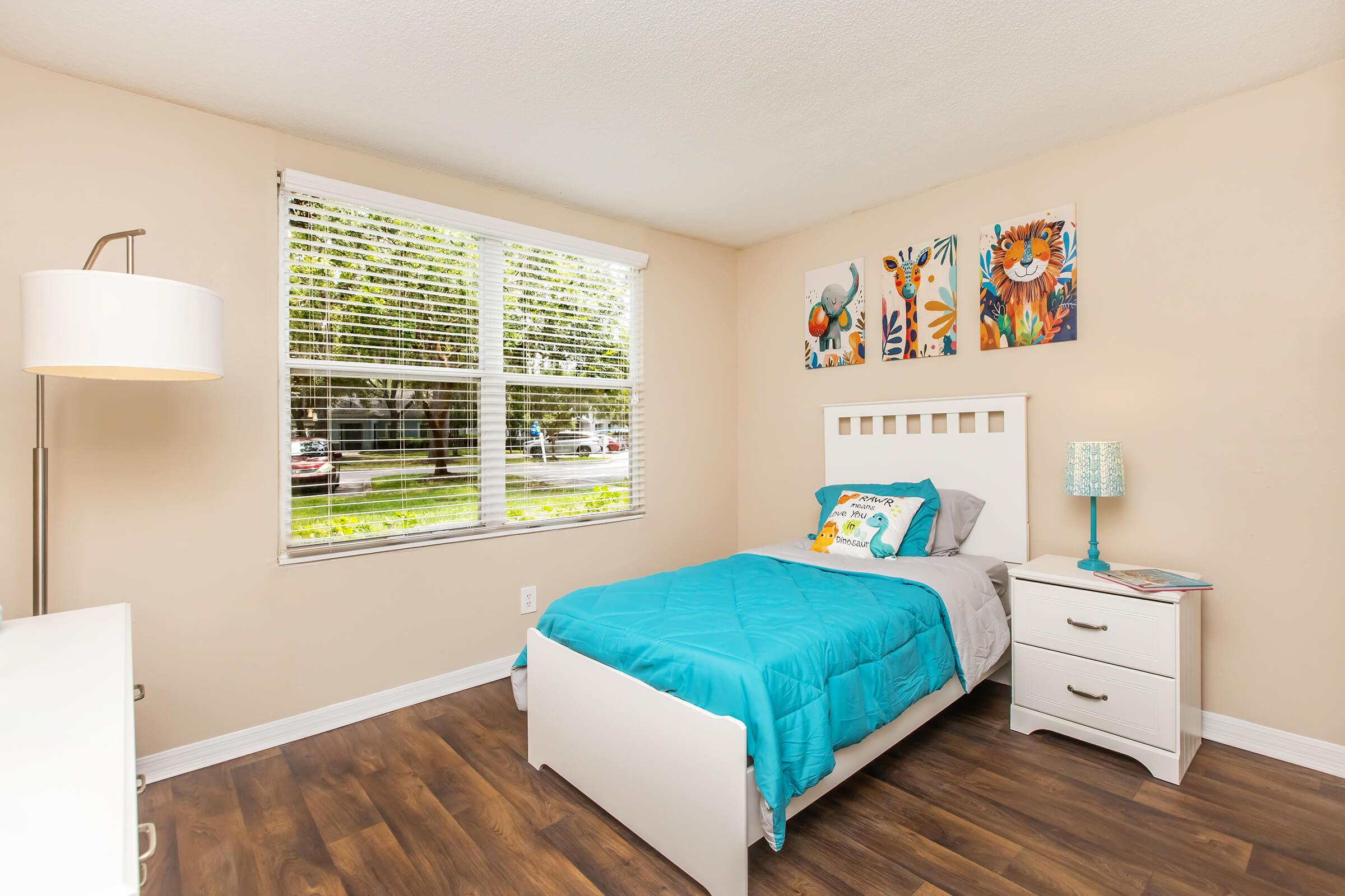 A cozy bedroom featuring a single bed with a turquoise quilt, a white nightstand with a lamp, and decorative artwork on the wall. Natural light streams through a window with blinds, illuminating the room's neutral walls and wooden flooring.