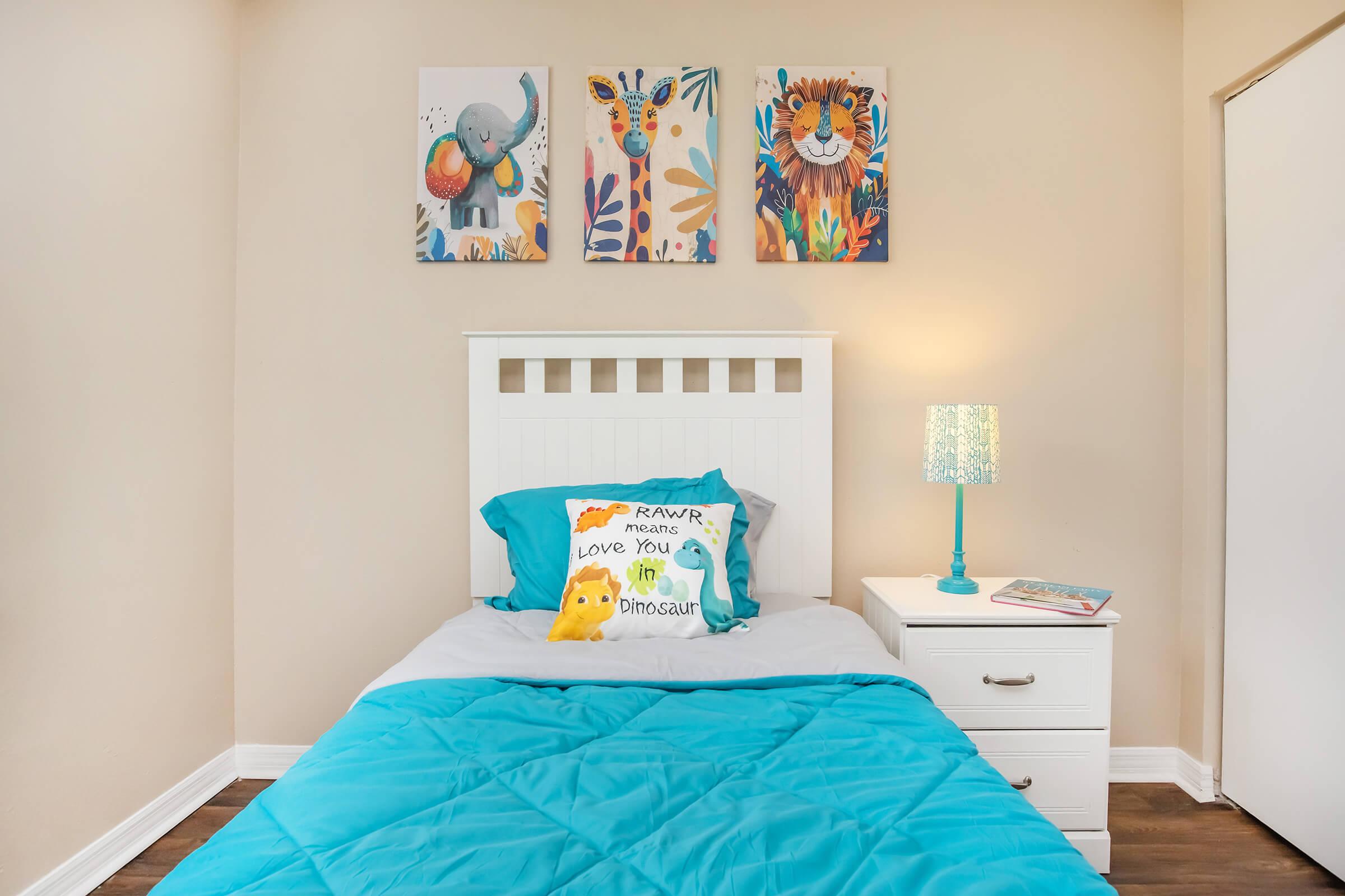 Cozy bedroom featuring a white bed with a turquoise comforter, decorated with a colorful dinosaur pillow. A bedside table holds a small lamp with a turquoise base. On the wall, there are three animal-themed art pieces: an elephant, a giraffe, and a lion. The room has light-colored walls and wooden flooring.