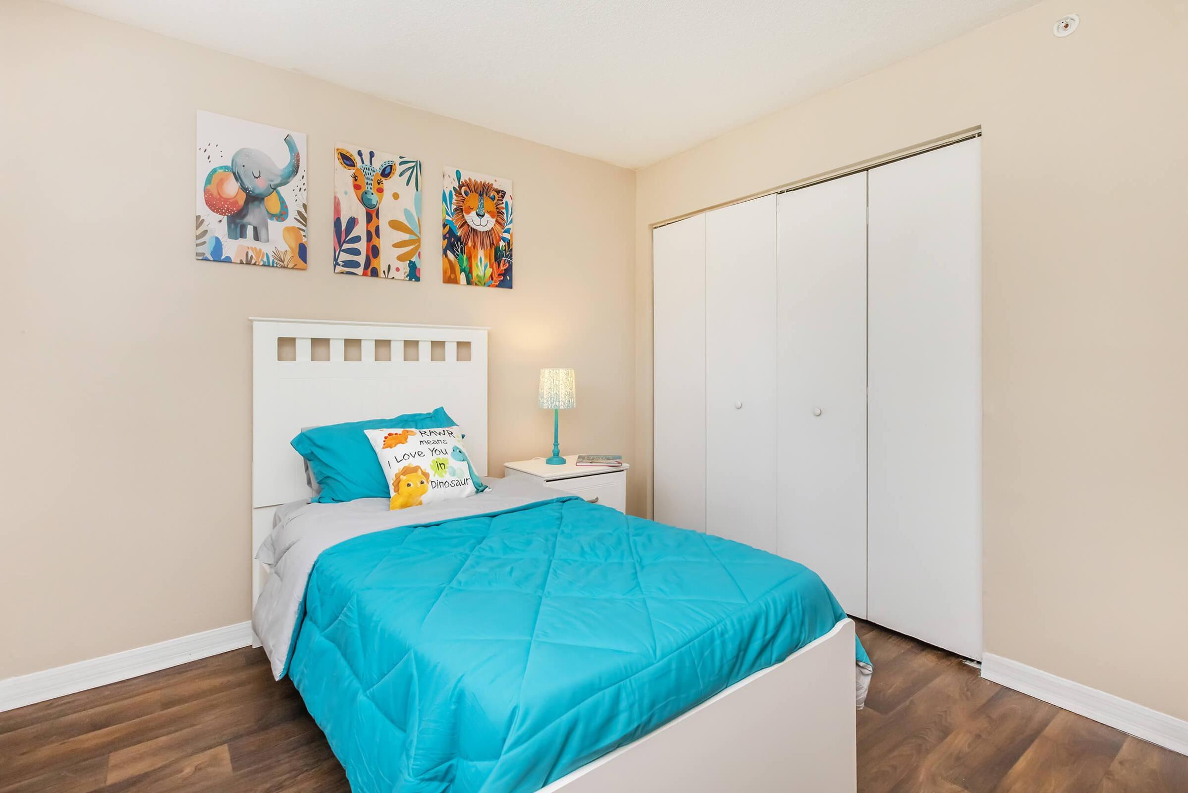 A cozy bedroom featuring a light blue comforter on a bed with decorative pillows. There are colorful animal-themed art pieces on the wall above the bed. A lamp on a nightstand adds soft lighting. The room has a warm beige wall color and white sliding closet doors, with wooden flooring creating a welcoming atmosphere.