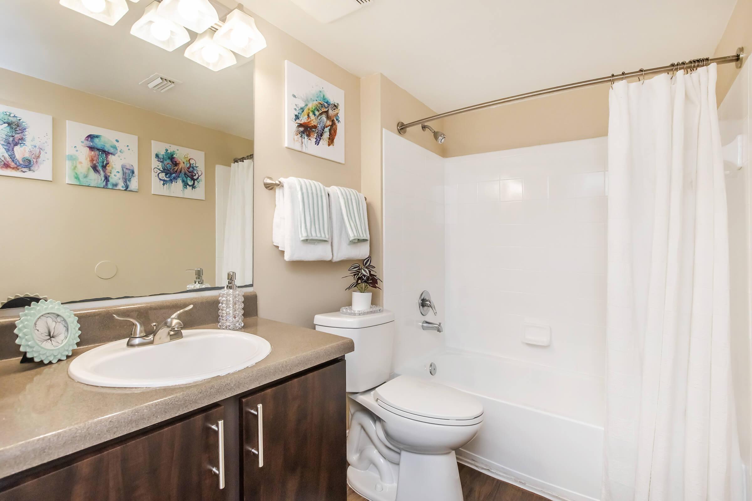 A modern bathroom featuring a sink with a round mirror, a white toilet, and a tub with a shower curtain. The walls are adorned with colorful art prints, and there are neatly arranged towels hanging on a bar. The decor includes a small potted plant and a decorative clock on the counter.