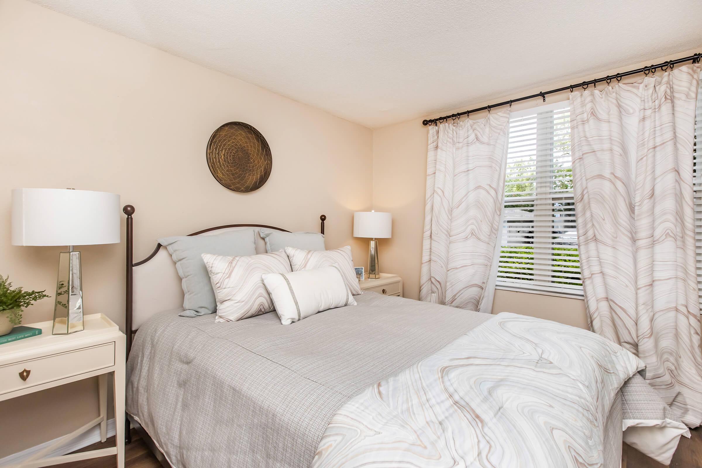 A cozy bedroom featuring a neatly made bed with a light gray and white patterned duvet. Two bedside tables with lamps flank the bed. A decorative wall plate hangs above the headboard, and sheer curtains partially cover a window, allowing natural light to illuminate the room. Greenery adds a touch of color.