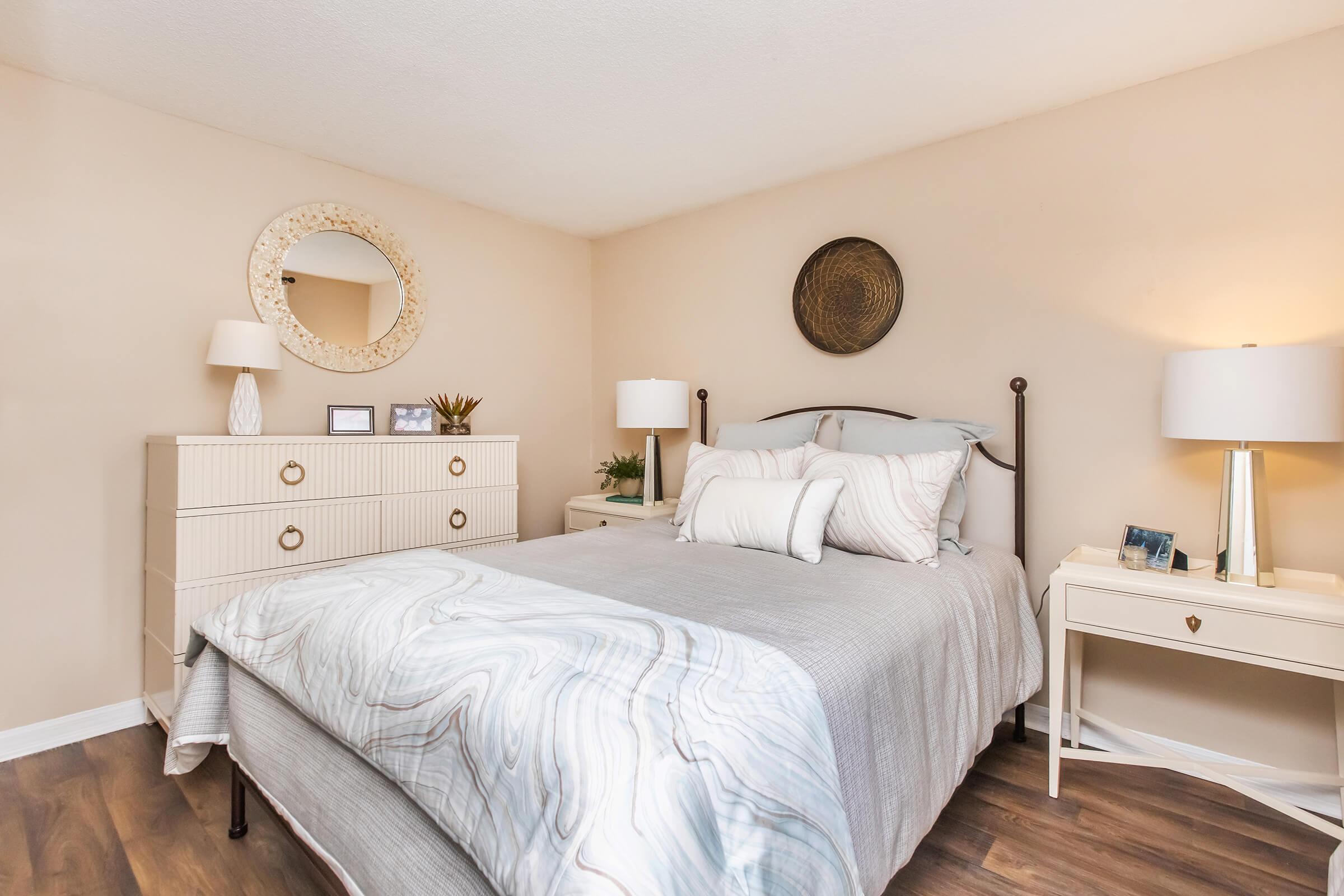 A cozy bedroom featuring a neatly made bed with light gray bedding, decorative pillows, and a stylish headboard. There's a white dresser with a round mirror above it, along with two bedside lamps on matching nightstands. Light tones and wood flooring create a warm, inviting atmosphere.