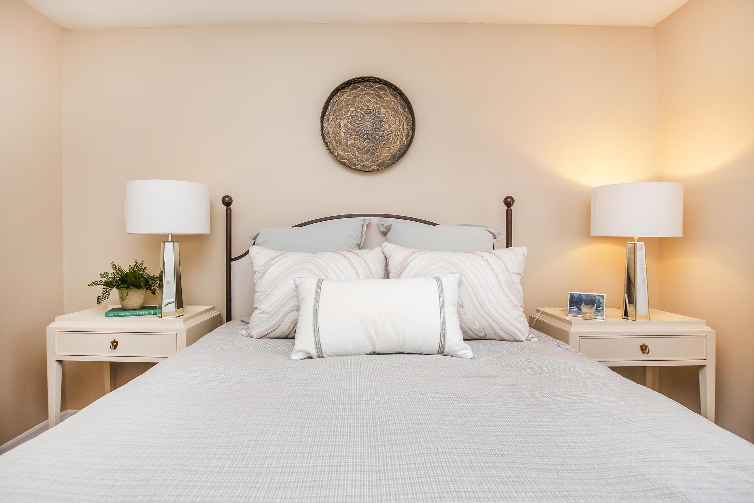 A cozy bedroom featuring a neatly made bed with light gray bedding, two bedside tables with lamps, and decorative pillows. A woven wall art piece is hung above the headboard, while a small plant and photo frame adorn the tables, creating a warm and inviting atmosphere.