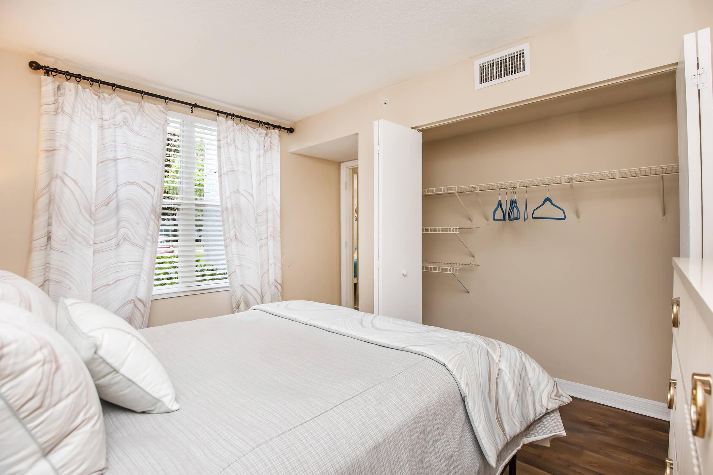 A cozy bedroom featuring a neatly made bed with light-colored bedding, decorative pillows, and large windows covered with sheer curtains. An open closet with wire shelving and a few blue hangers is visible, and the walls are painted a soft beige, creating a warm and inviting atmosphere.