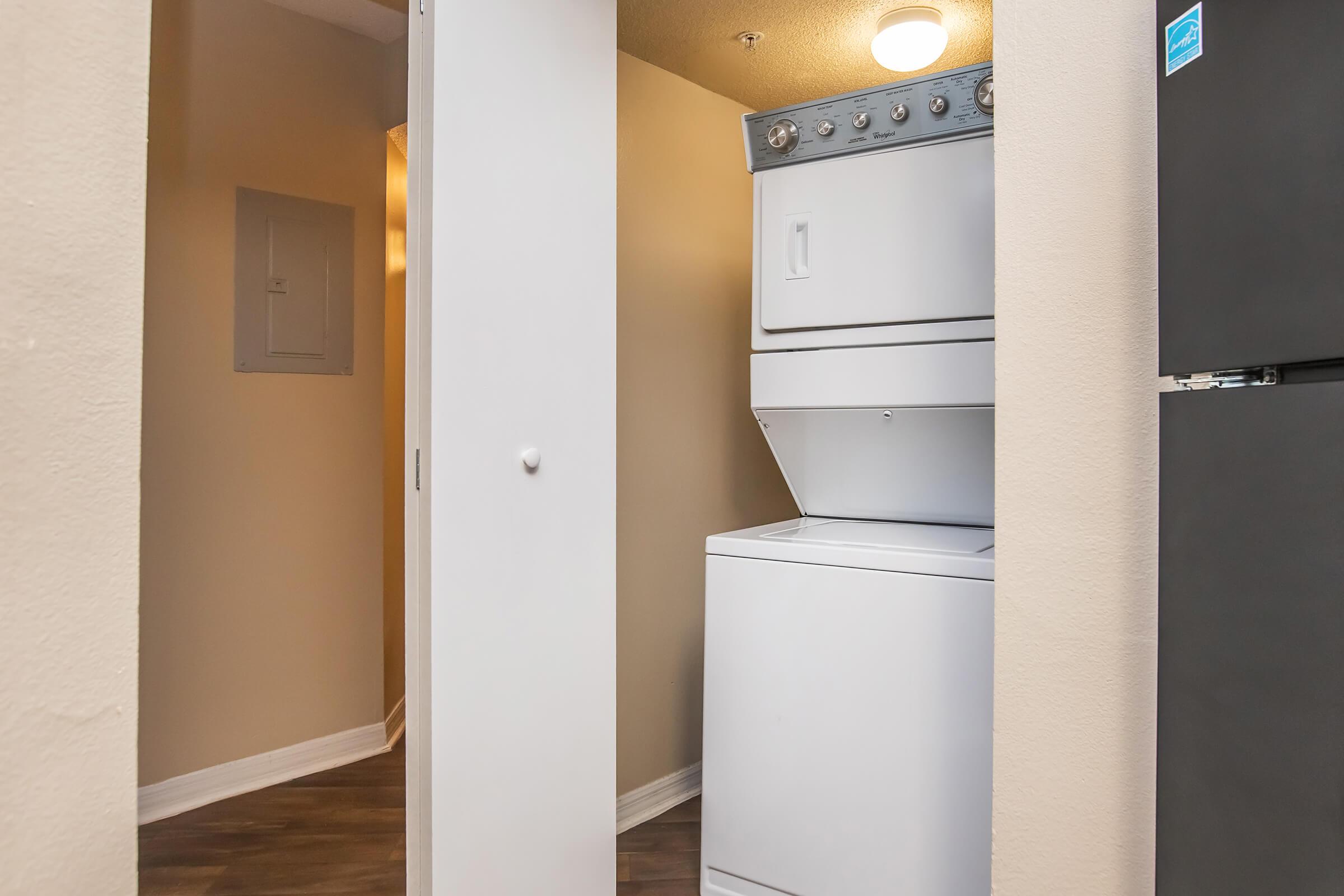 A compact laundry area featuring a stacked washer and dryer against a neutral-colored wall. The space is minimally decorated, with a doorway leading to another room in the background. A refrigerator can be seen partially on the right side of the image.
