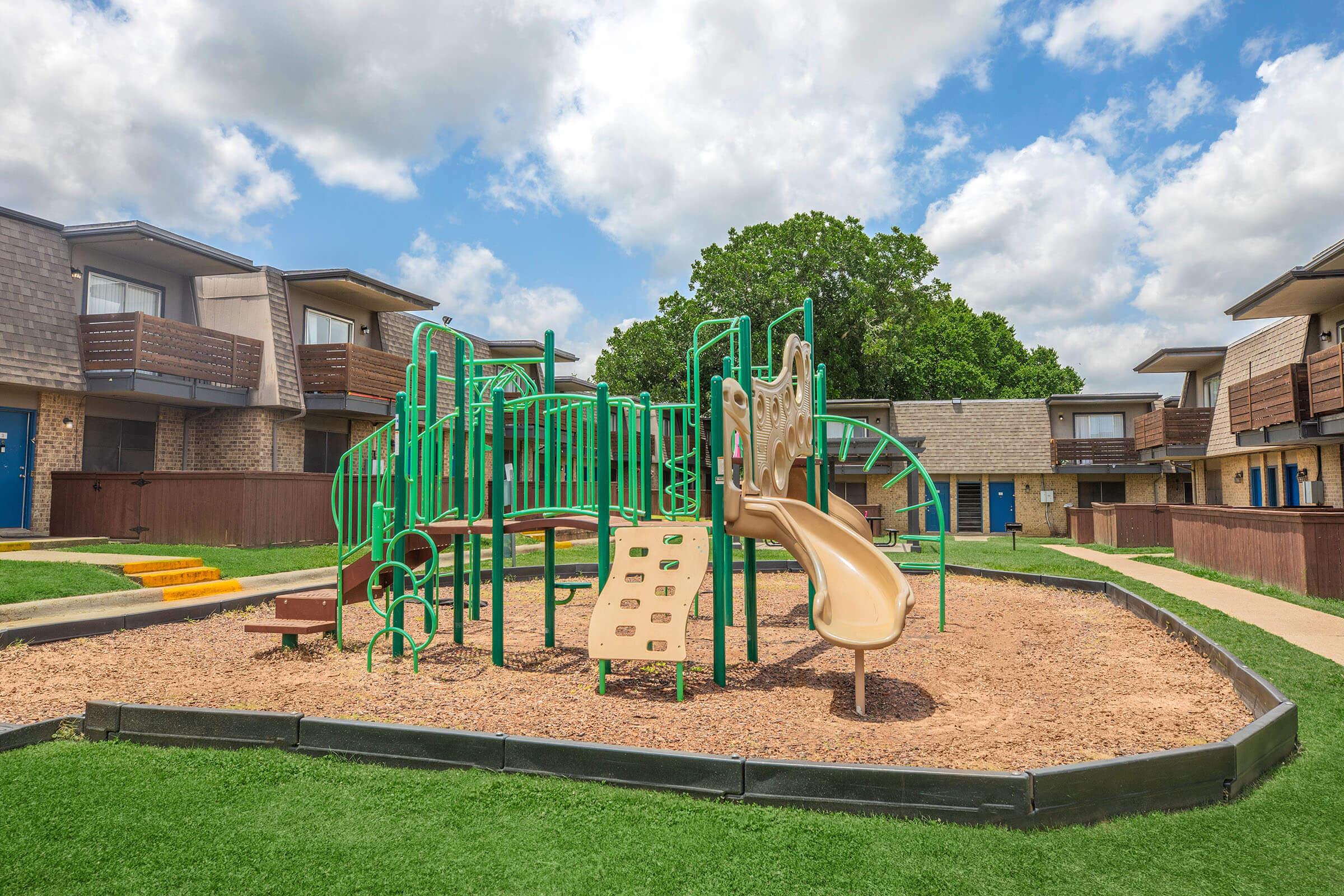 Playground area featuring green and brown play equipment, including slides, climbing structures, and a grassy surrounding. The background shows two-story apartment buildings with balconies and a blue sky with fluffy clouds.