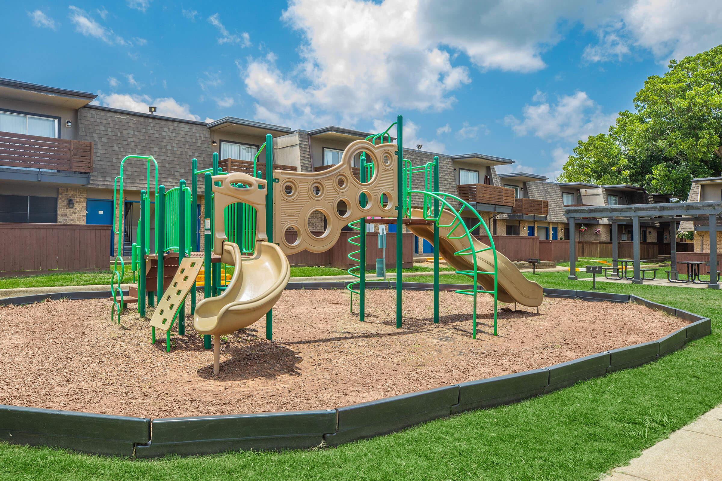 Playground with green and tan climbing structures, slides, and a spiral slide, surrounded by wood chip mulch. In the background, there are several apartment buildings with balconies and a bright blue sky with scattered clouds. Green grass surrounds the play area, enhancing the outdoor setting.