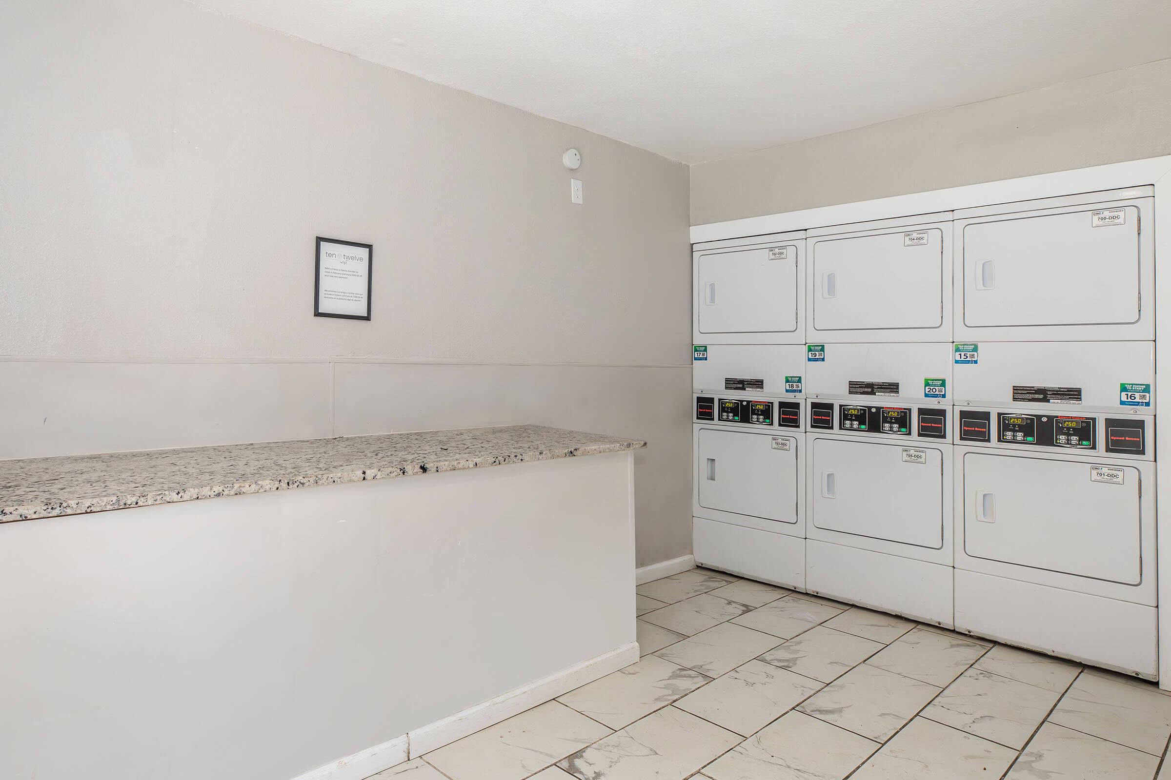 A clean and simple laundry room featuring a beige wall, a granite countertop, and several stacked washing machines and dryers with control panels. The floor is tiled, and there is a framed sign on the wall. The space is well-lit and organized, providing a functional area for laundry tasks.