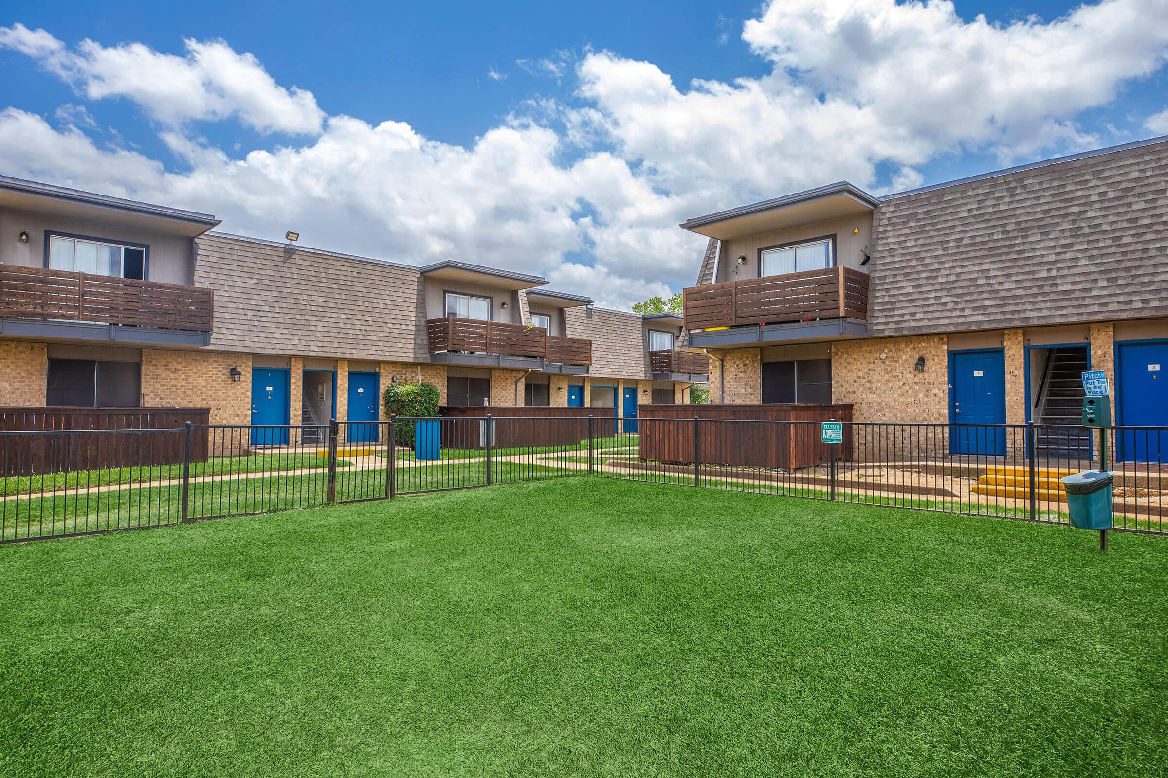 A view of a residential apartment complex featuring several two-story buildings with blue doors and wooden balconies. There is a green lawn area enclosed by a black fence, with a few small trees and shrubs. The sky is partly cloudy, creating a bright and inviting atmosphere.