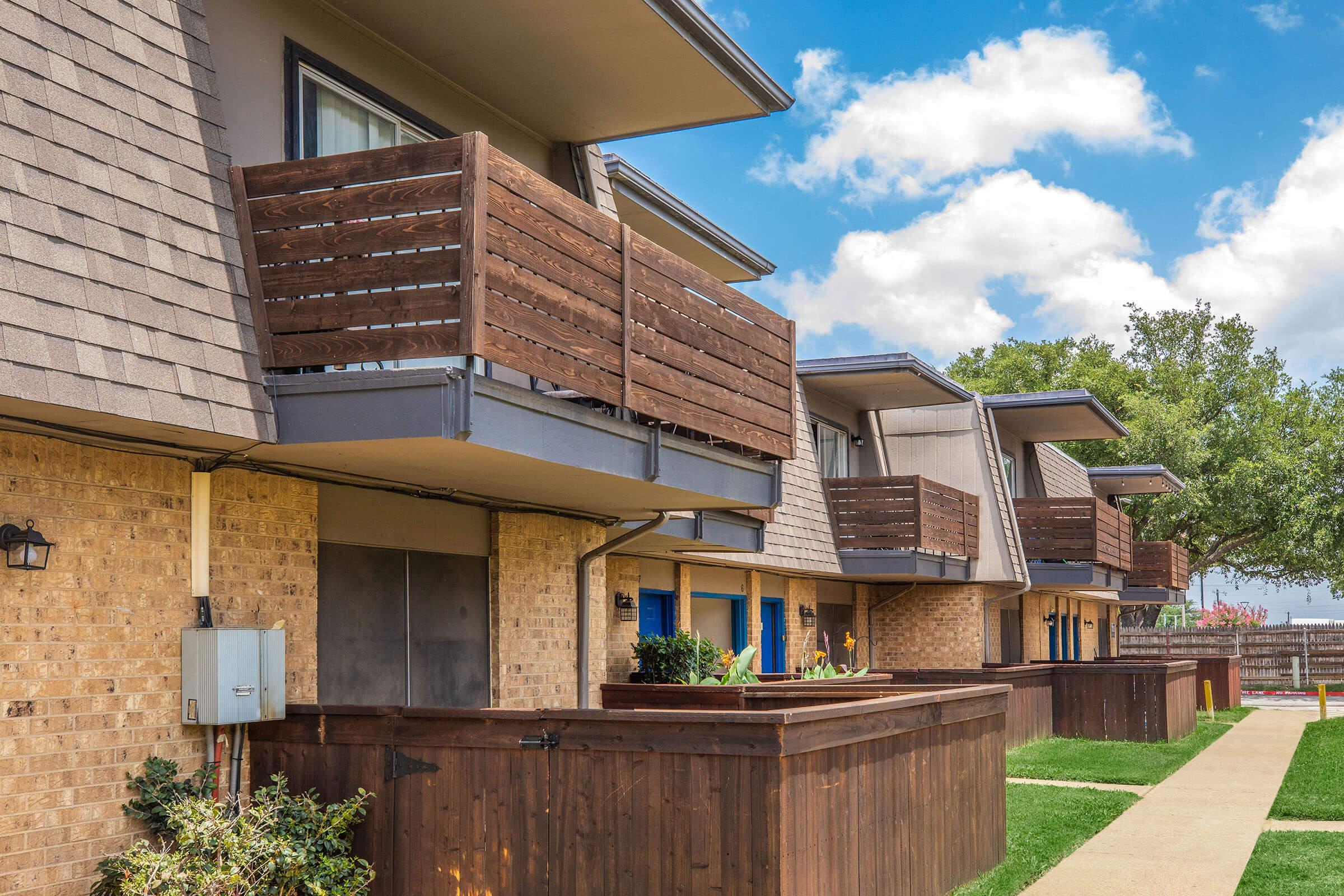 Brick building with modern design featuring multiple balconies made of wood. Green grass lines the walkway, which leads to the entrances. The sky is partly cloudy, and there are trees in the background, creating a pleasant residential atmosphere.