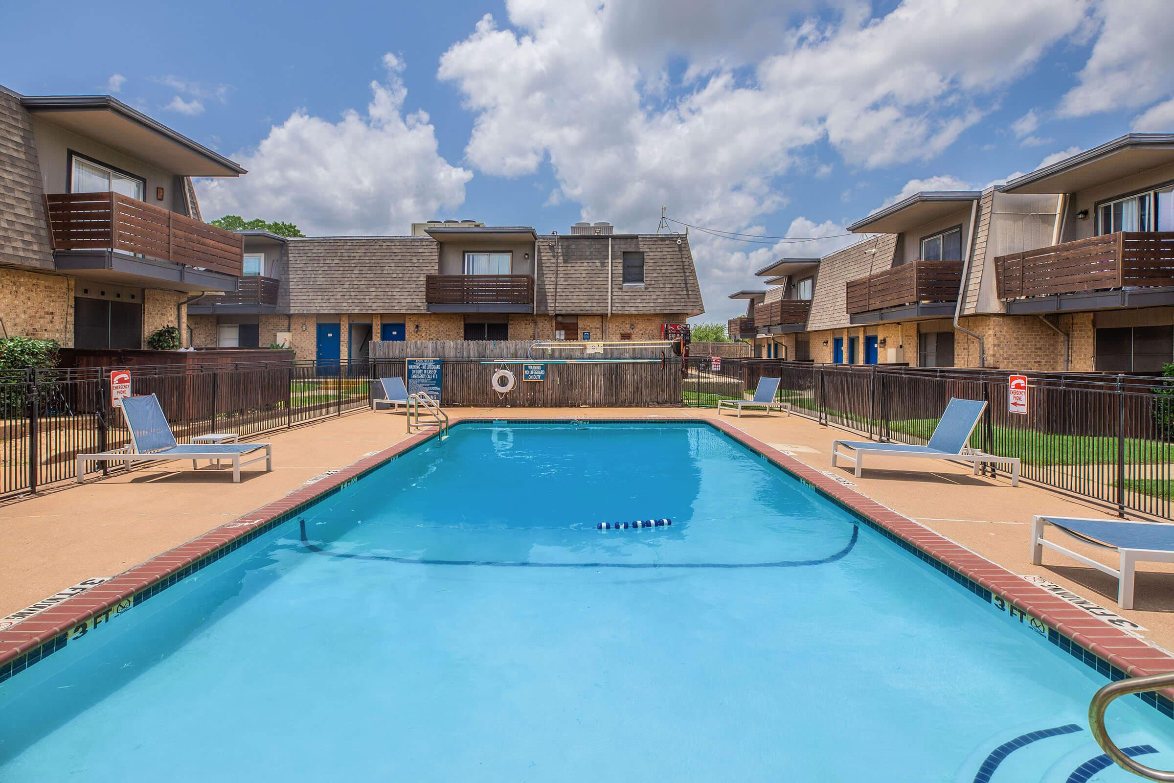 A clear blue swimming pool surrounded by lounge chairs, with two-story apartment buildings in the background. The sky is partly cloudy, and a fence encloses the pool area.