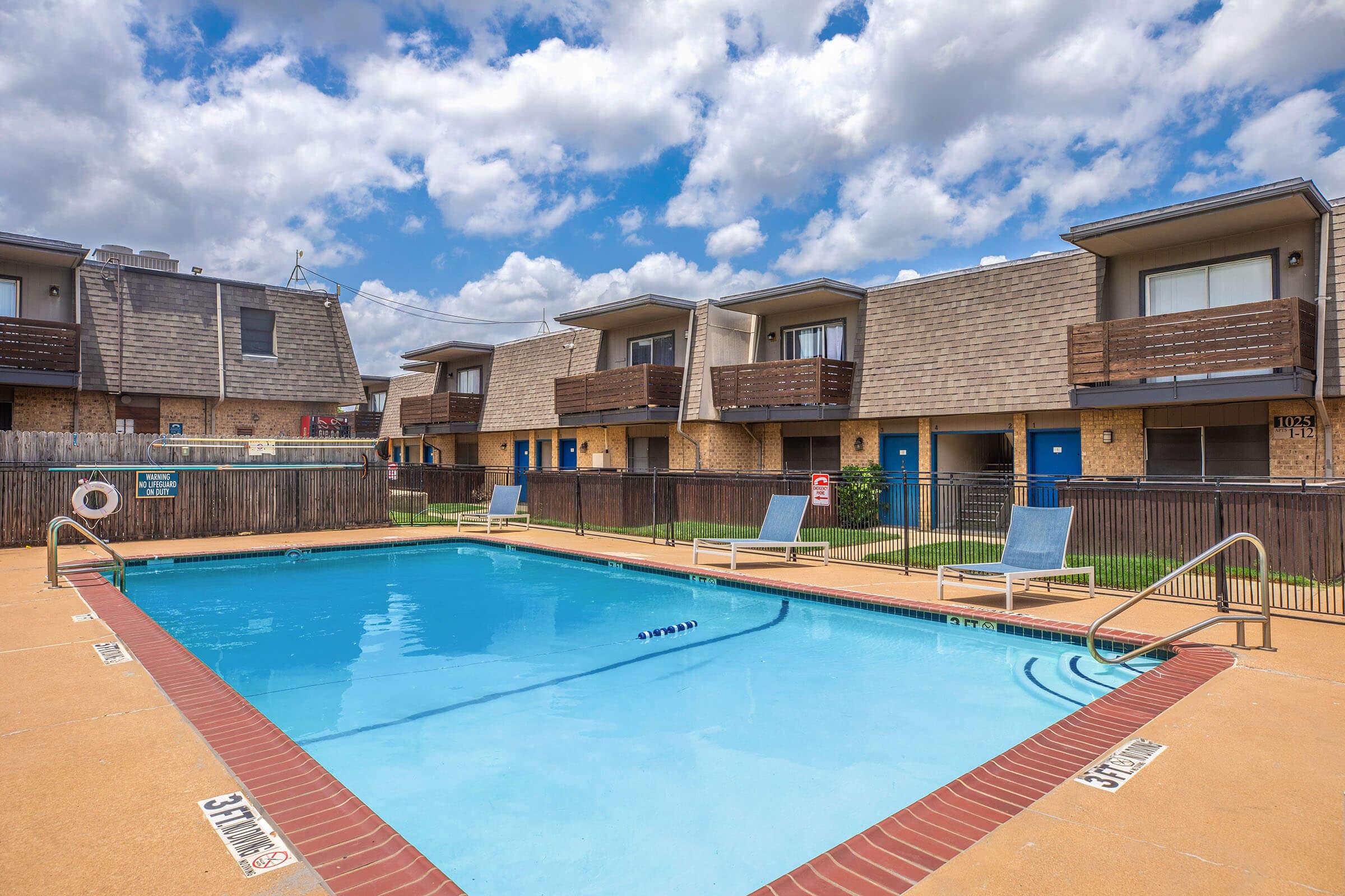 A clear blue swimming pool surrounded by a tan deck area, with lounge chairs on the side. In the background, there are two-story apartment buildings featuring balconies and blue doors, set against a partly cloudy sky.