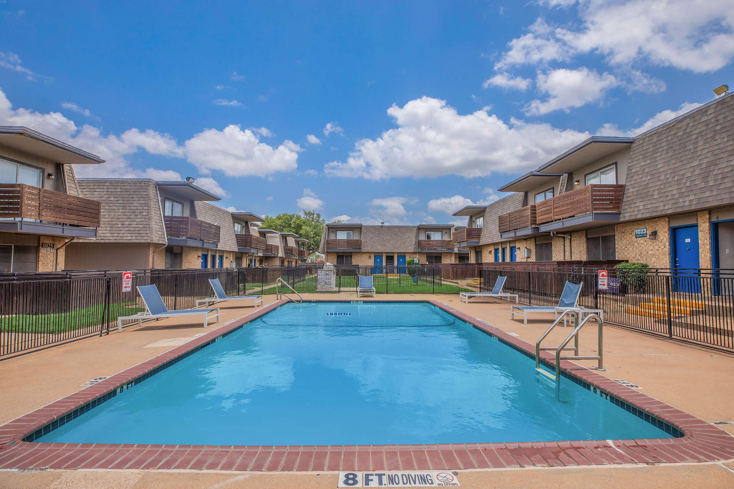 A clear blue swimming pool surrounded by lounge chairs, with two-story apartments in the background. The area is sunny with a few clouds in the sky and marked with an "8 FT. NO DIVING" sign near the pool's edge. Lush green grass lines the pool area, enhancing the summer vibe.