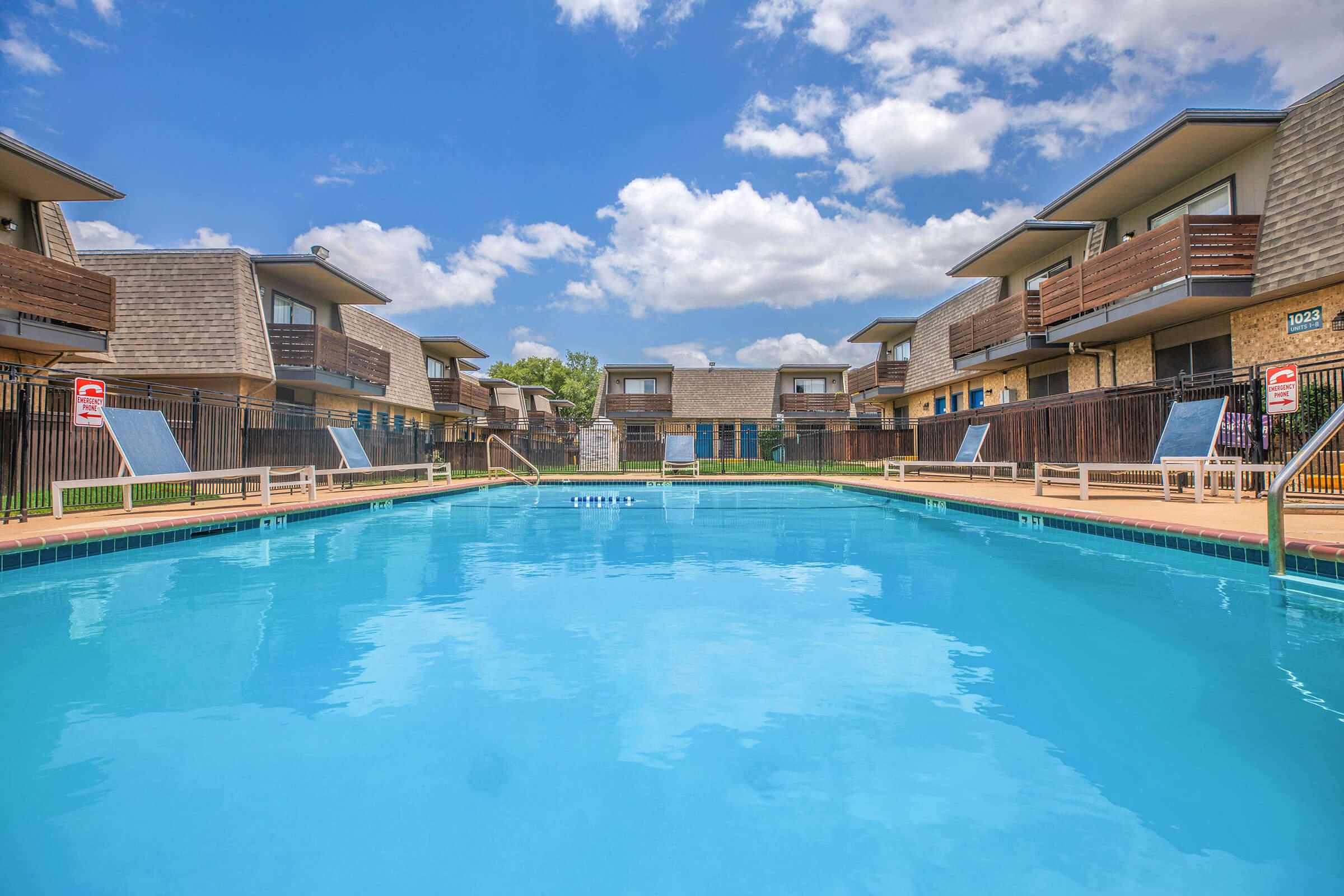 A clear blue swimming pool surrounded by brown residential buildings. The pool area features lounge chairs and is bordered by a fence. The sky is bright with scattered clouds, creating a sunny and inviting atmosphere ideal for relaxation.