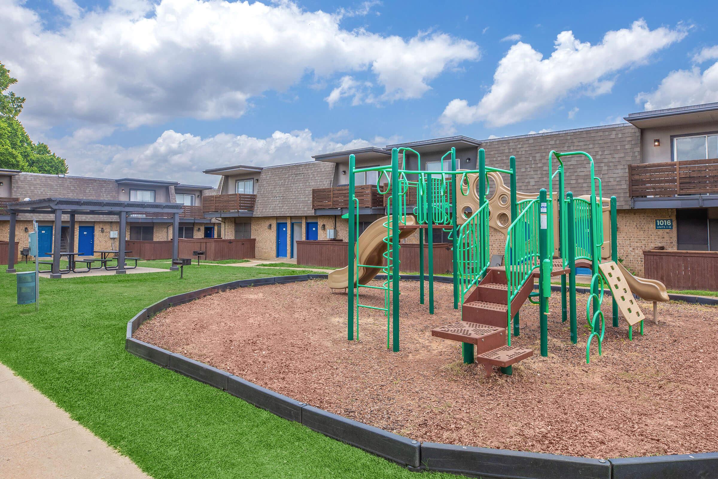 Playground area featuring green climbing structures, slides, and a sandbox, surrounded by grassy areas. In the background, there are several residential buildings with blue doors. The sky is partly cloudy, creating a bright and inviting atmosphere.