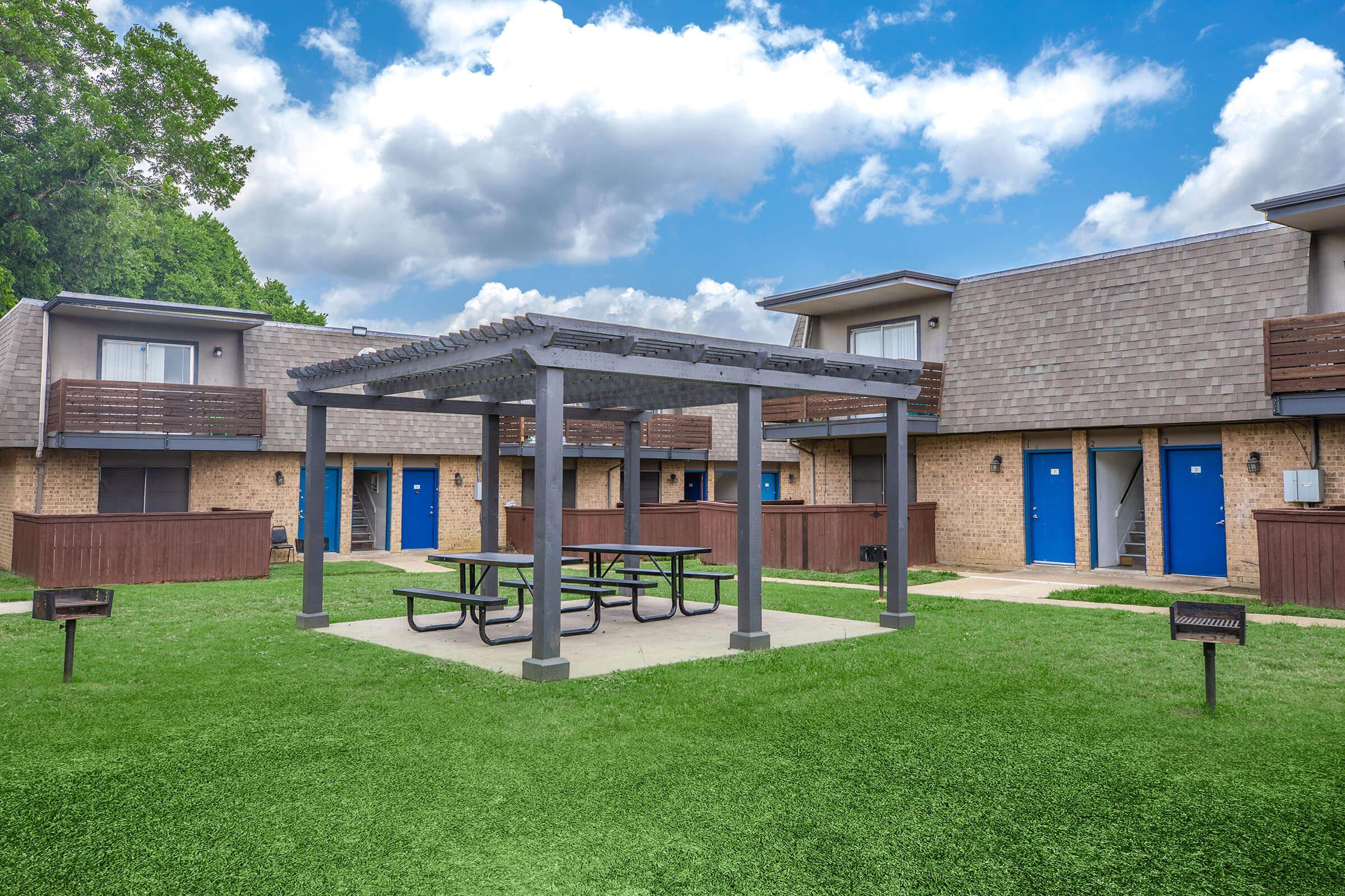A courtyard area featuring a picnic table under a wooden pergola, surrounded by green lawns. Brown brick buildings with blue doors are visible in the background, along with additional picnic and grilling areas. The sky is partly cloudy, creating a bright and inviting atmosphere.