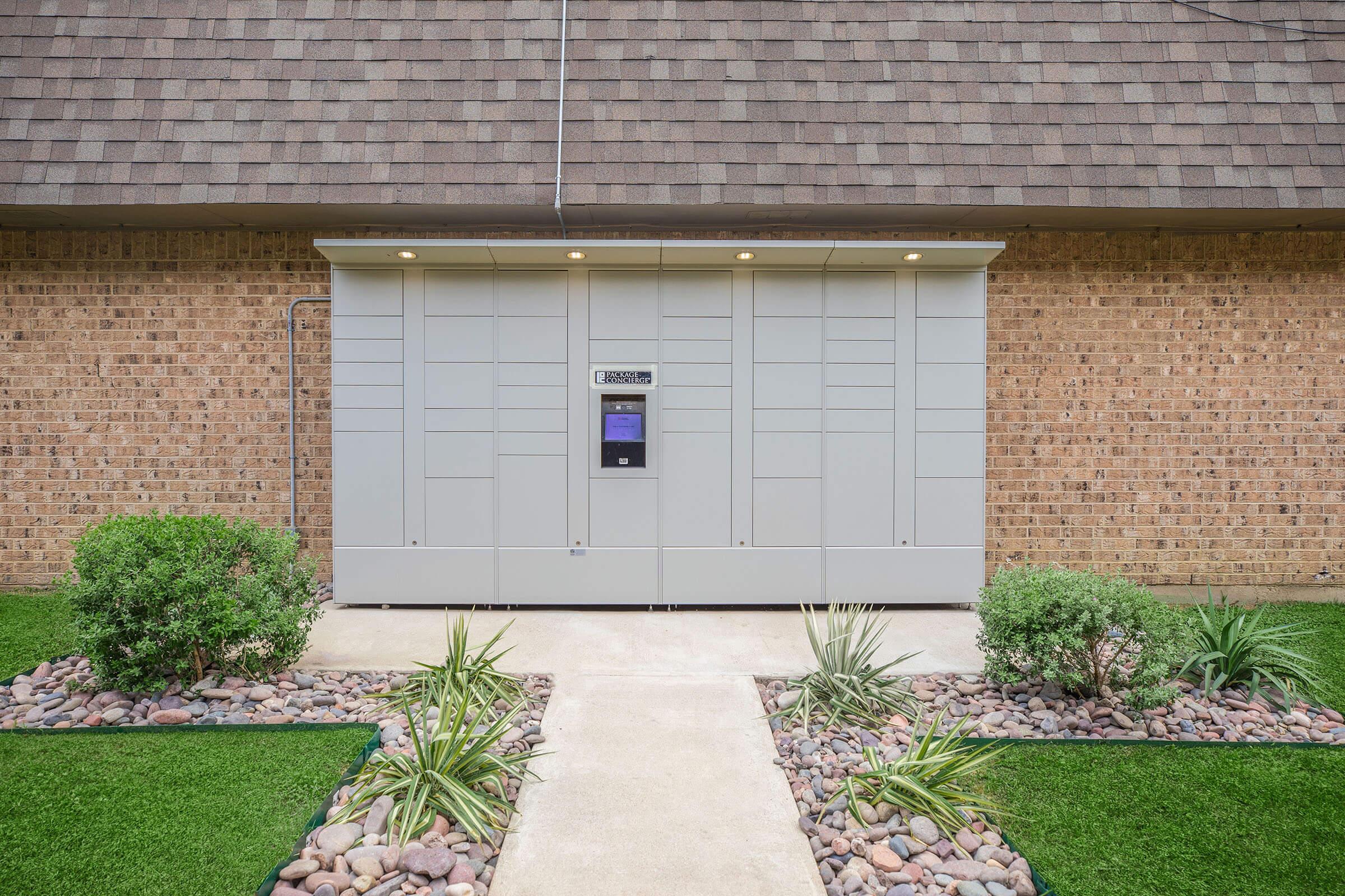 A modern gray delivery box mounted against a brick wall, with a digital screen and buttons. The surrounding area features a neatly landscaped path lined with small rocks and low shrubs, leading up to the delivery box. Soft lighting is installed above the box for visibility.
