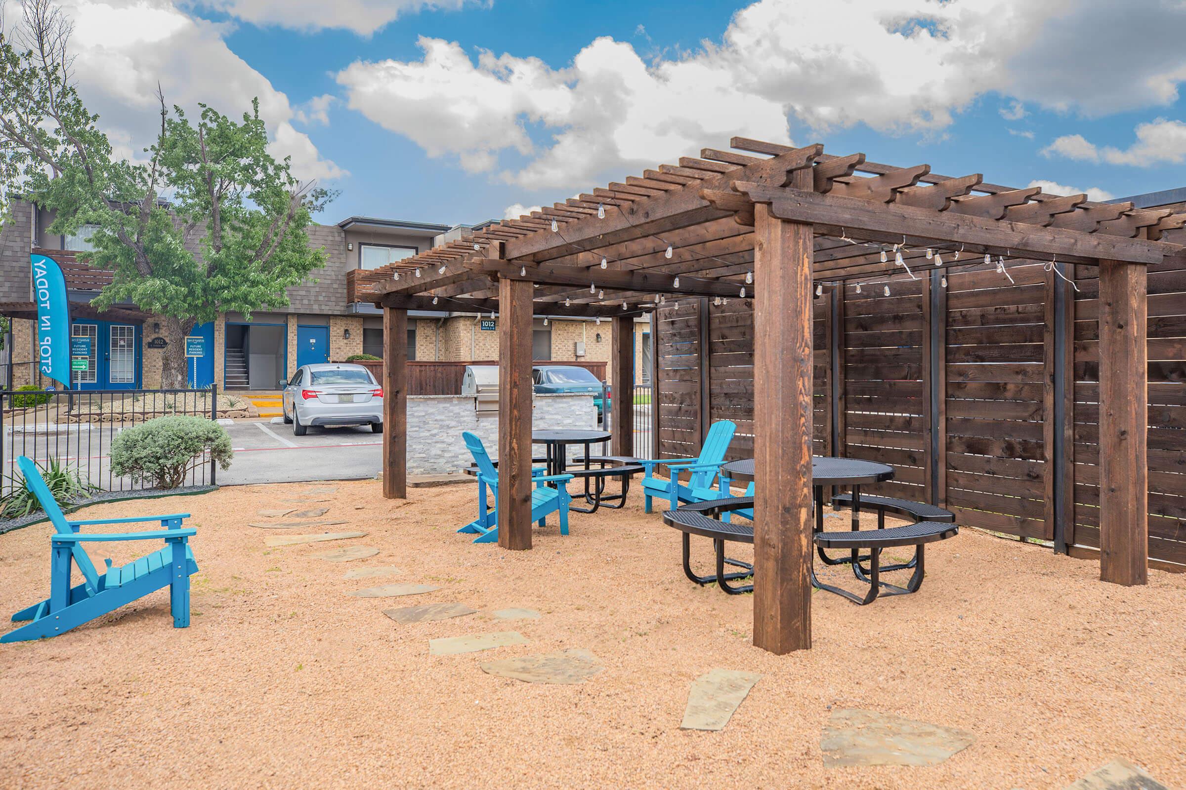 A cozy outdoor seating area featuring a wooden pergola with hanging lights, surrounded by a gravel landscape. Brightly colored blue adirondack chairs complement a black picnic table. In the background, there are nearby buildings and green trees under a partly cloudy sky.