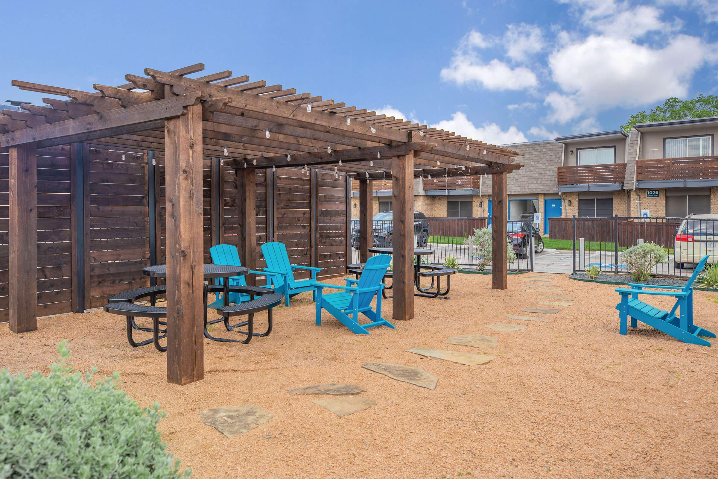 Outdoor communal area featuring a wooden pergola and turquoise Adirondack chairs. Gravel surface with stone pathways leads to a seating arrangement, creating a cozy atmosphere. In the background, there are two-story buildings and a parking area. Bright blue sky with a few clouds above.