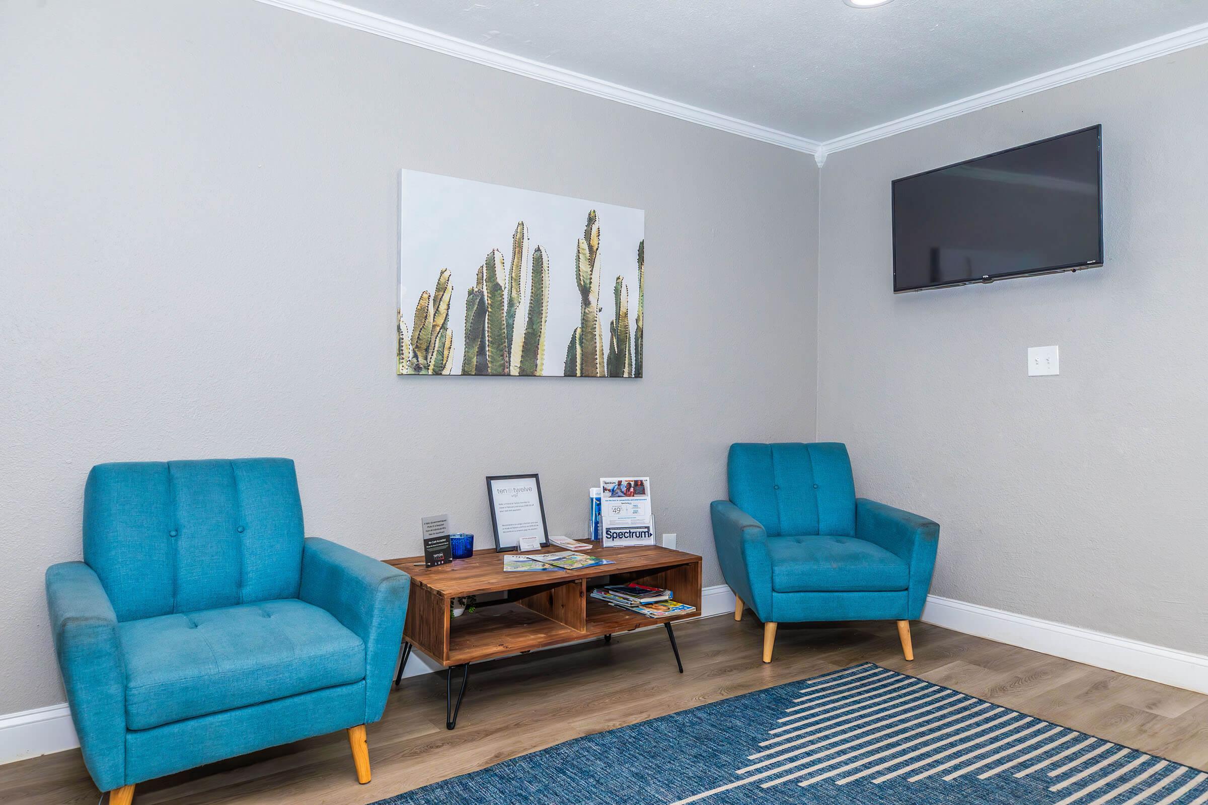 A modern waiting area featuring two turquoise chairs, a wooden coffee table with magazines, and a decorative wall art of cacti. There is a flat-screen TV mounted on the wall and a blue patterned rug on the floor, all set against a neutral gray background.