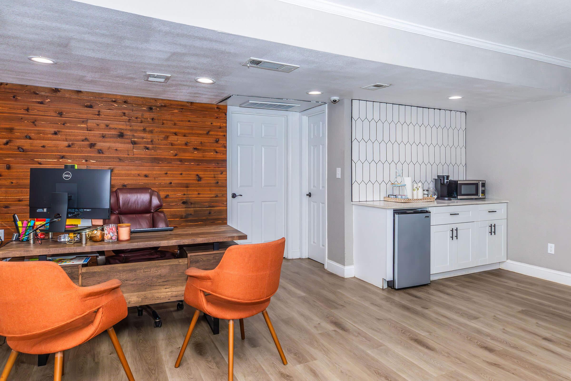 A modern office space featuring a wooden accent wall, a desk with a computer, and two orange chairs. There's a kitchenette with a microwave, mini-fridge, and cabinets, set against a light-colored wall. The flooring is wood-like, creating a warm and inviting atmosphere.