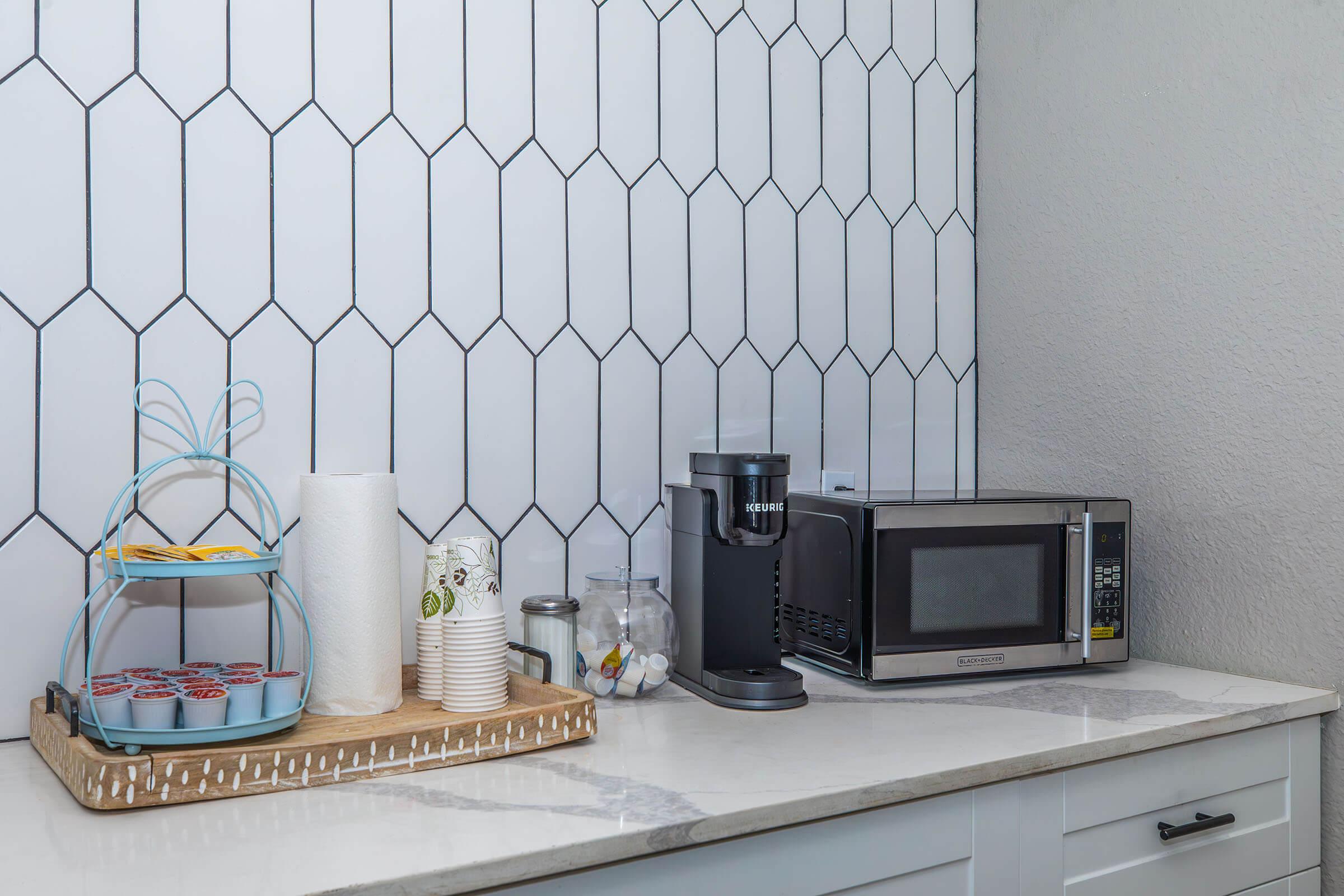 A modern kitchen countertop featuring a hexagonal tiled backsplash. On the left, a tiered metal stand holds baked goods. To the right, there is a roll of paper towels, disposable cups, a glass jar, a microwave, and a coffee maker, all set against a clean, bright countertop.