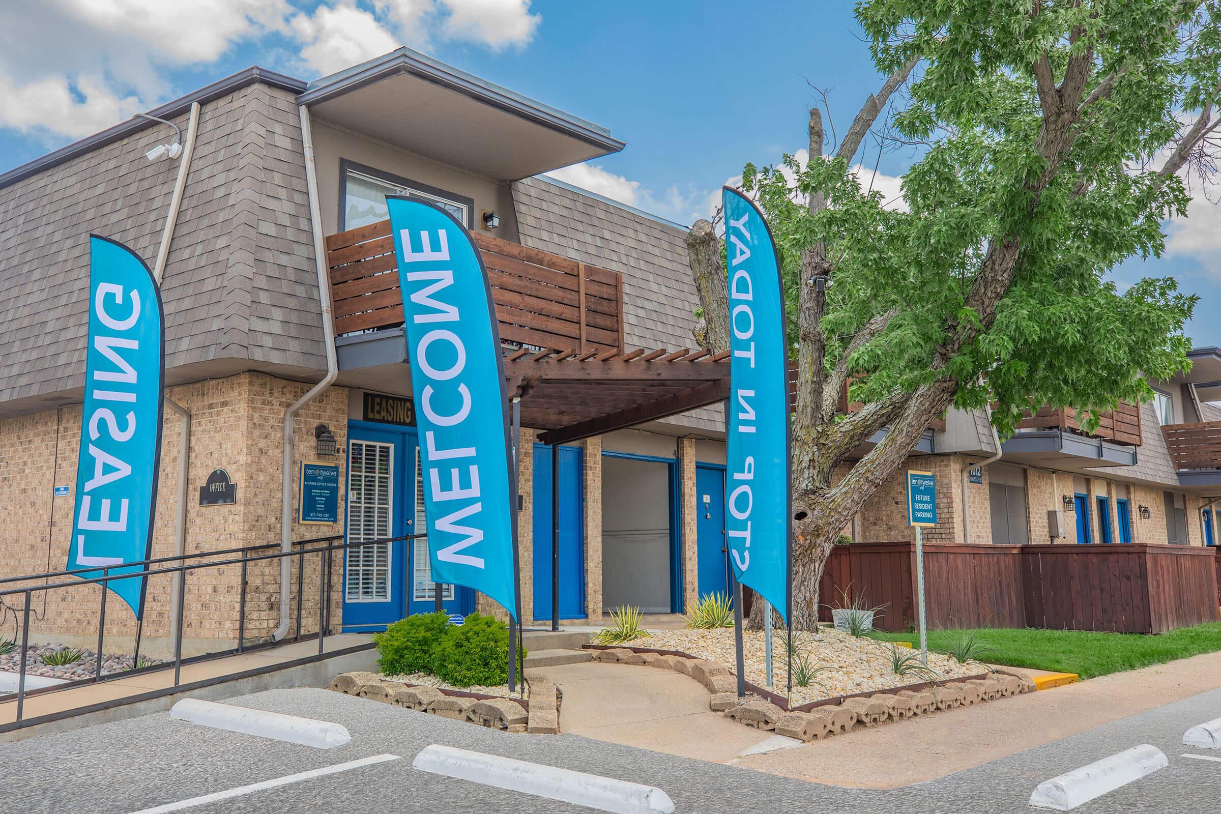 A leasing office with blue flags that read "Welcome" and "Stop in Today!" The building features a modern design, stone exterior, and a landscaped area with trees. A ramp leads to the entrance, and there are parking spaces in front. The sky is partly cloudy, creating a bright atmosphere.
