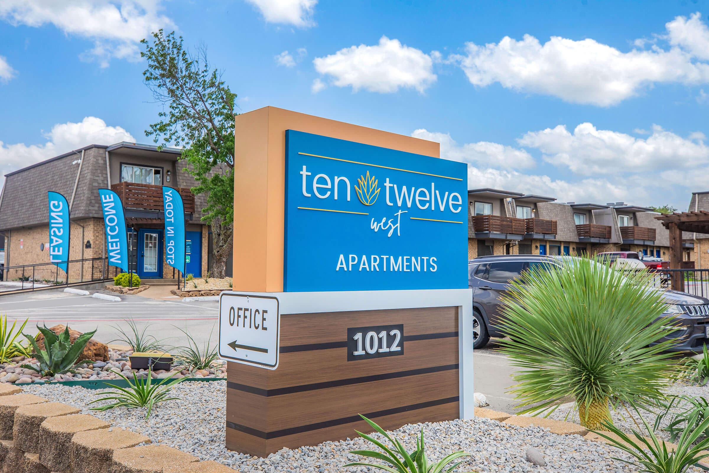 Sign for Ten Twelve West Apartments, featuring a blue and orange design. The sign includes the text "Ten Twelve West Apartments" and directional arrows to the office. The background shows a well-maintained landscape with a palm-like plant and a clear blue sky with clouds.