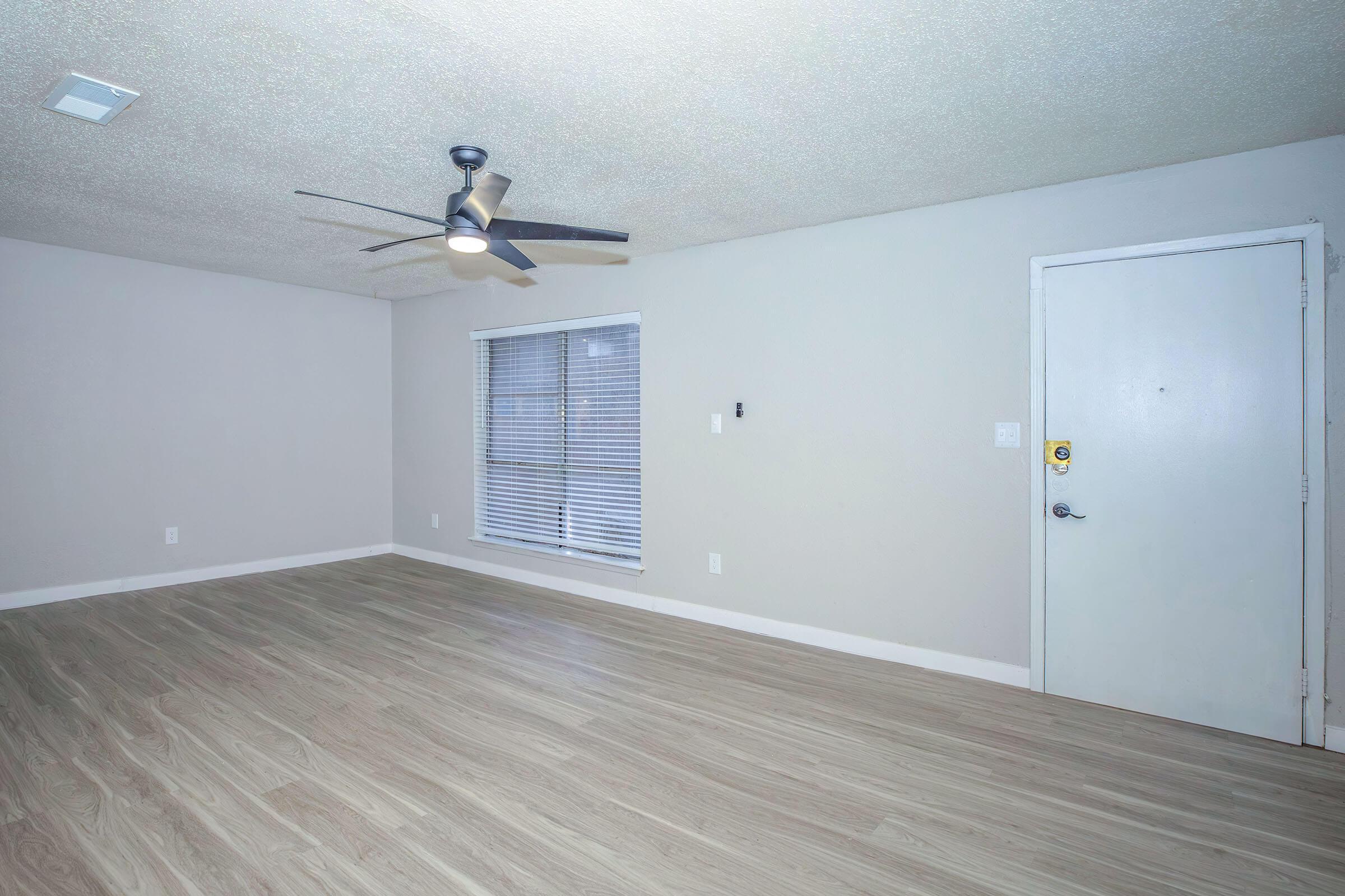A spacious, empty room with light-colored walls and a ceiling fan. The floor is wood-like, and there is a window with blinds allowing natural light. A door is visible on the right side, and the room has a clean, modern look, ideal for personal decoration or use.