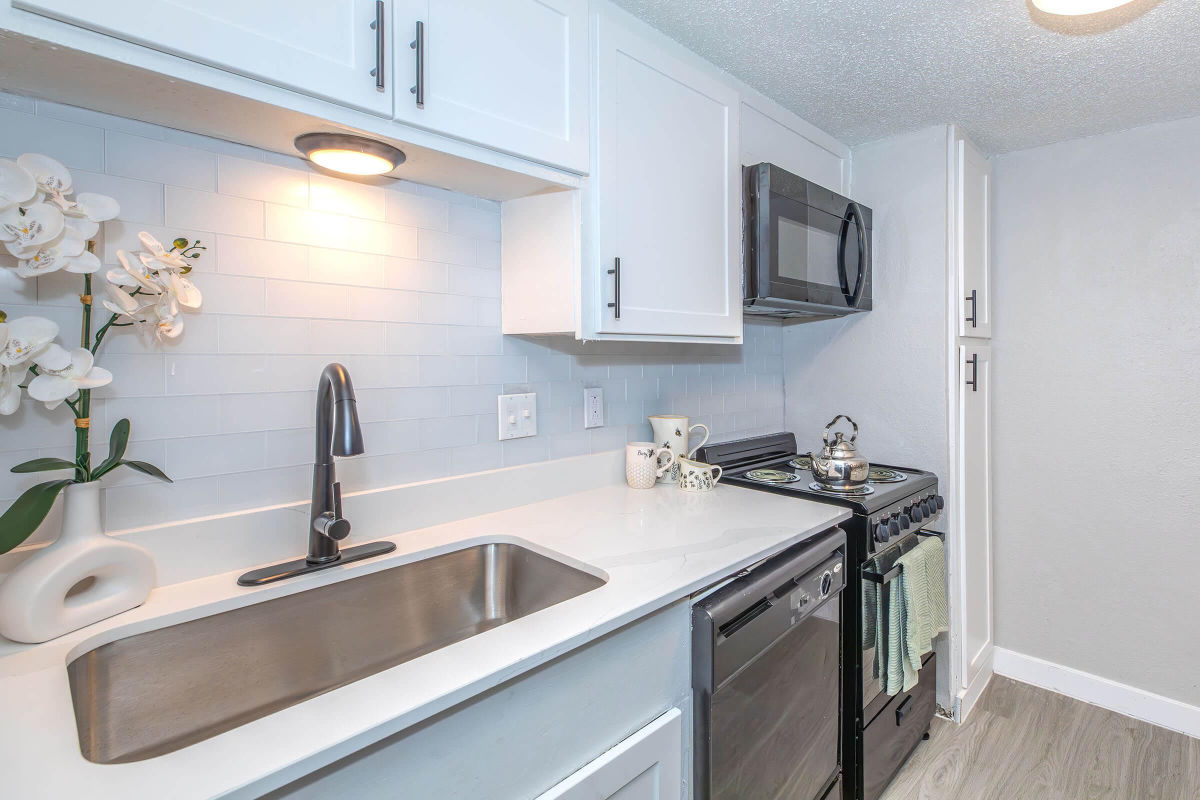 Modern kitchen with white cabinets, stainless steel appliances, and a sleek sink. A potted orchid is placed on the countertop, adding a touch of elegance. The backsplash features light-colored tiles, and dish towels hang from the oven, creating a warm and inviting atmosphere.
