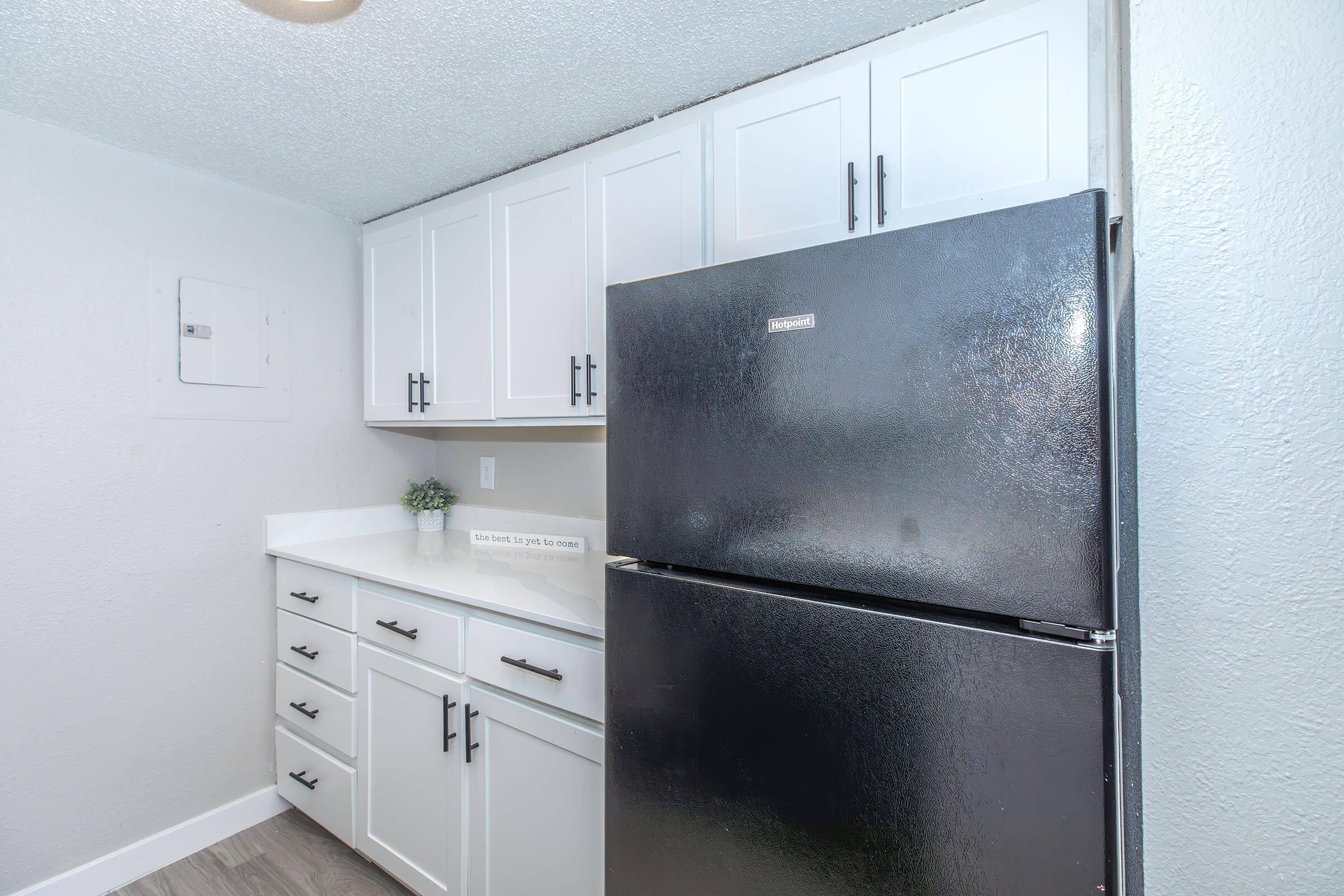 A modern kitchen featuring white cabinets with black handles, a black refrigerator, and a light-colored countertop. The walls are painted in a soft shade, and there is a small decorative plant on the countertop. The space is well-lit and designed for functionality.