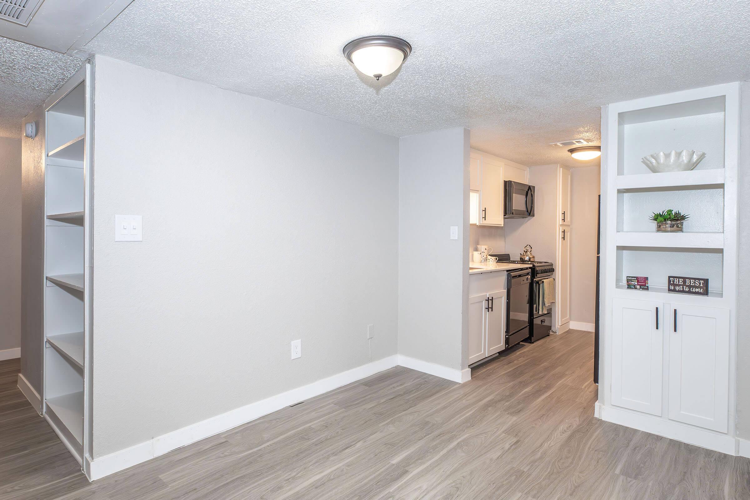 Interior view of a modern kitchen and living area in an apartment. The kitchen features white cabinets, stainless steel appliances, and a small counter space. Light gray walls and flooring give a contemporary feel. A built-in bookshelf is visible on the right side, adding storage and decor options.