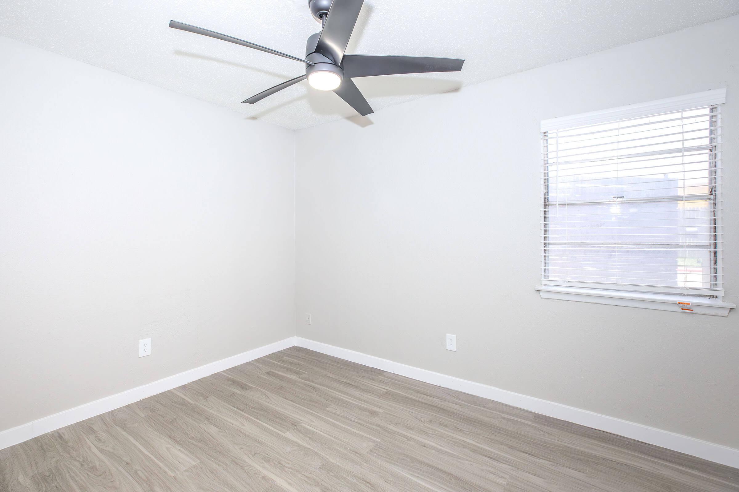 Interior view of a sparsely furnished room featuring light gray walls, a ceiling fan with five blades, and a window with horizontal blinds. The room has a light wood-like floor and white trim, creating a clean and modern appearance.