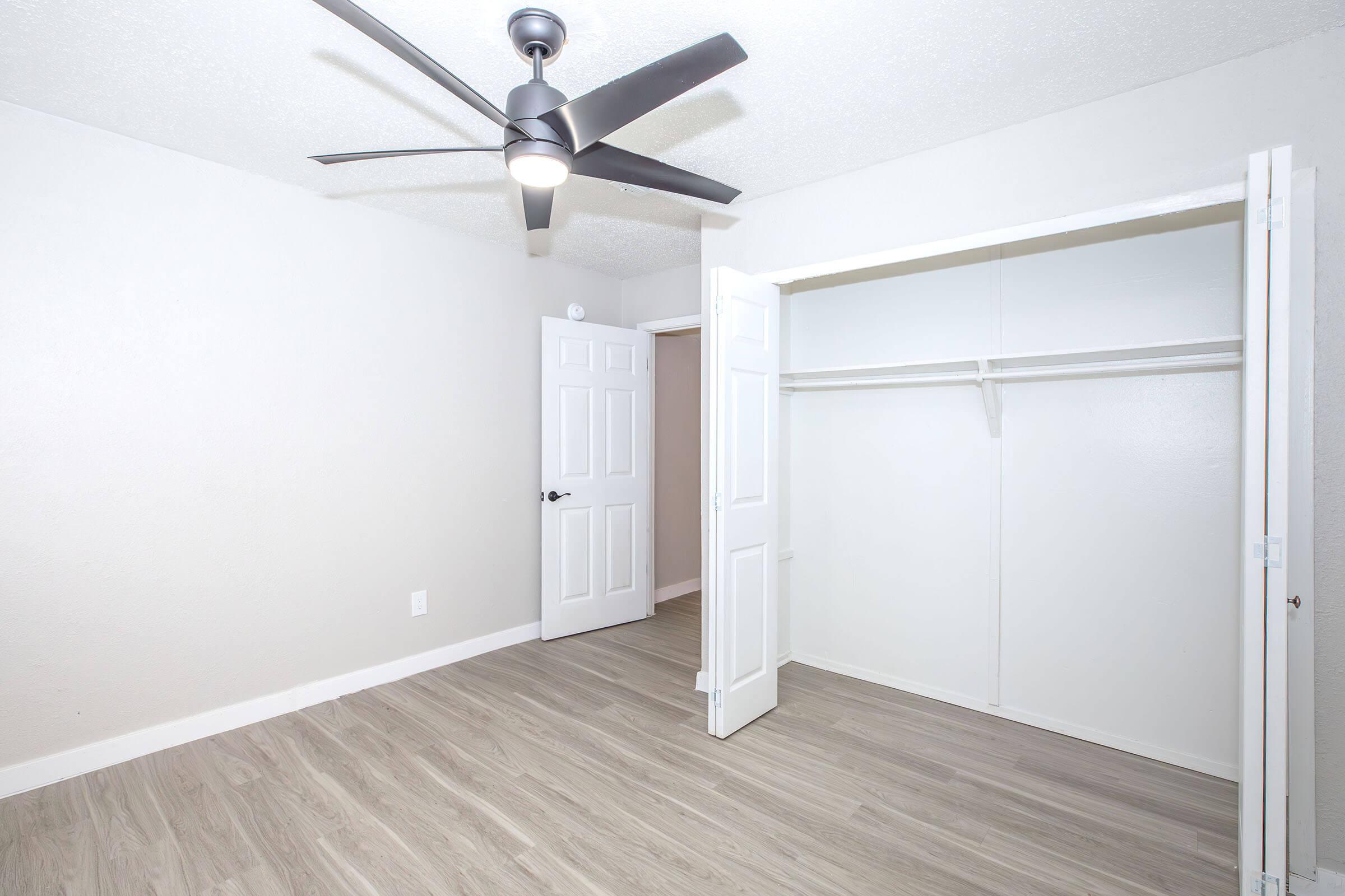 A well-lit, empty room featuring a light-colored wall, a ceiling fan, and a closet with open double doors. The flooring is a light wood-like material, and there is a door leading to another area, enhancing the spacious and clean appearance of the room.