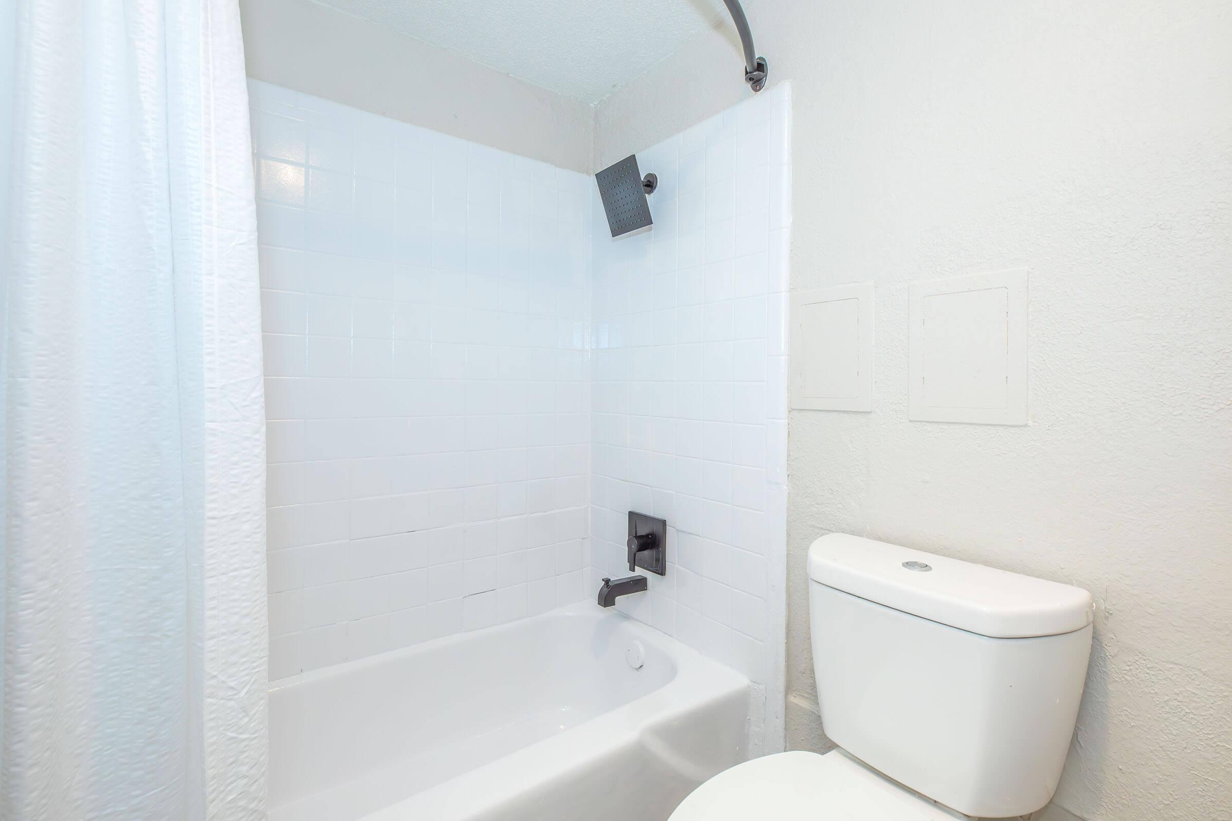 A clean and minimalistic bathroom featuring a white bathtub and a modern showerhead. The walls are tiled with white ceramic tiles, and there is a white toilet nearby. The overall aesthetic is bright and simple, emphasizing a contemporary design.