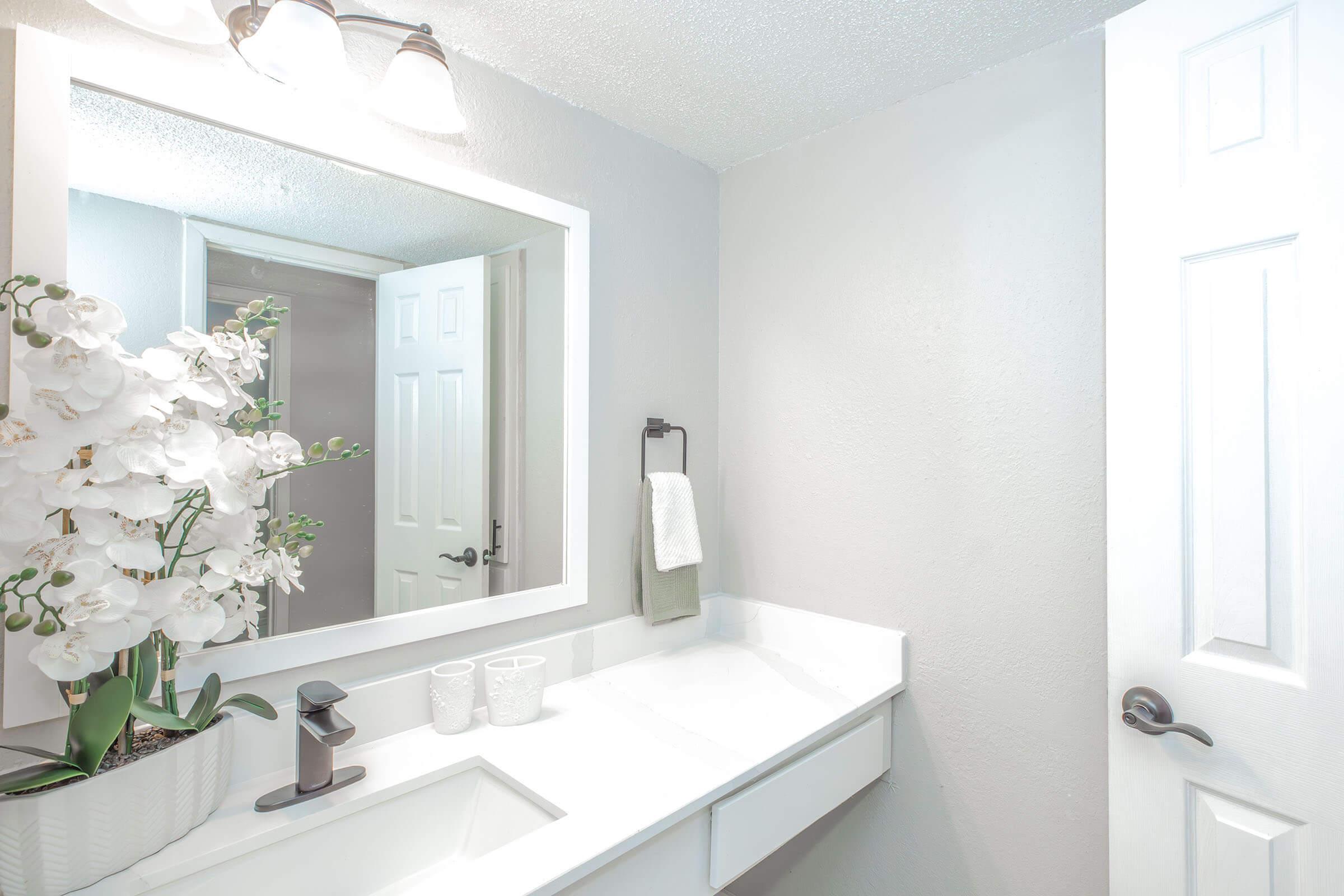 A modern bathroom featuring a white countertop and sink, a large mirror with backlighting, and decorative white orchids. The walls are painted light gray, and there is a towel holder with a towel. A closed door is visible on the right, adding to the clean and minimalist aesthetic of the space.