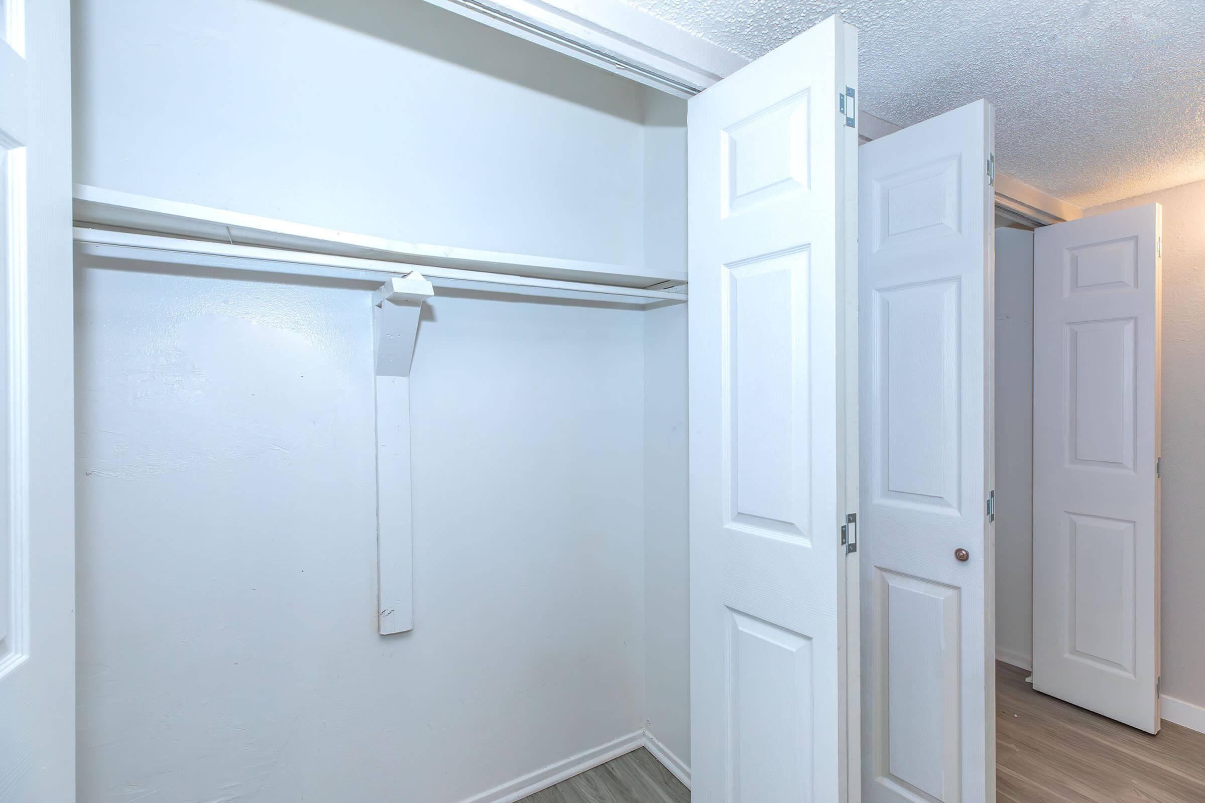 Empty closet with white walls and a hanging rod. The closet has multiple white doors, two of which are slightly open, revealing the interior space. The flooring appears light-colored and modern.