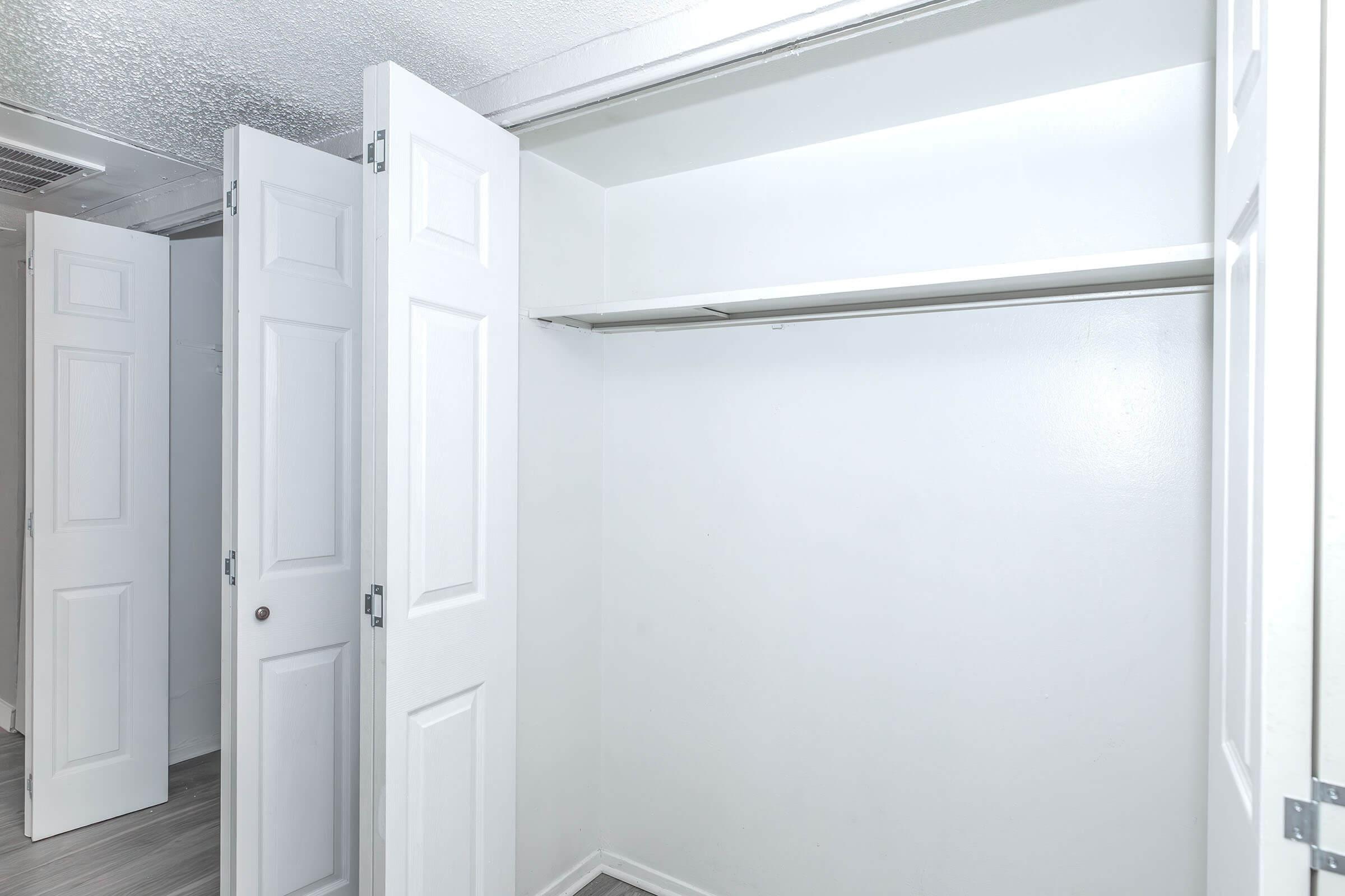 An empty white closet with two sets of double doors open, revealing a bare shelf along the top and clean walls. The flooring is light-colored, enhancing the spacious and minimalist appearance of the closet.