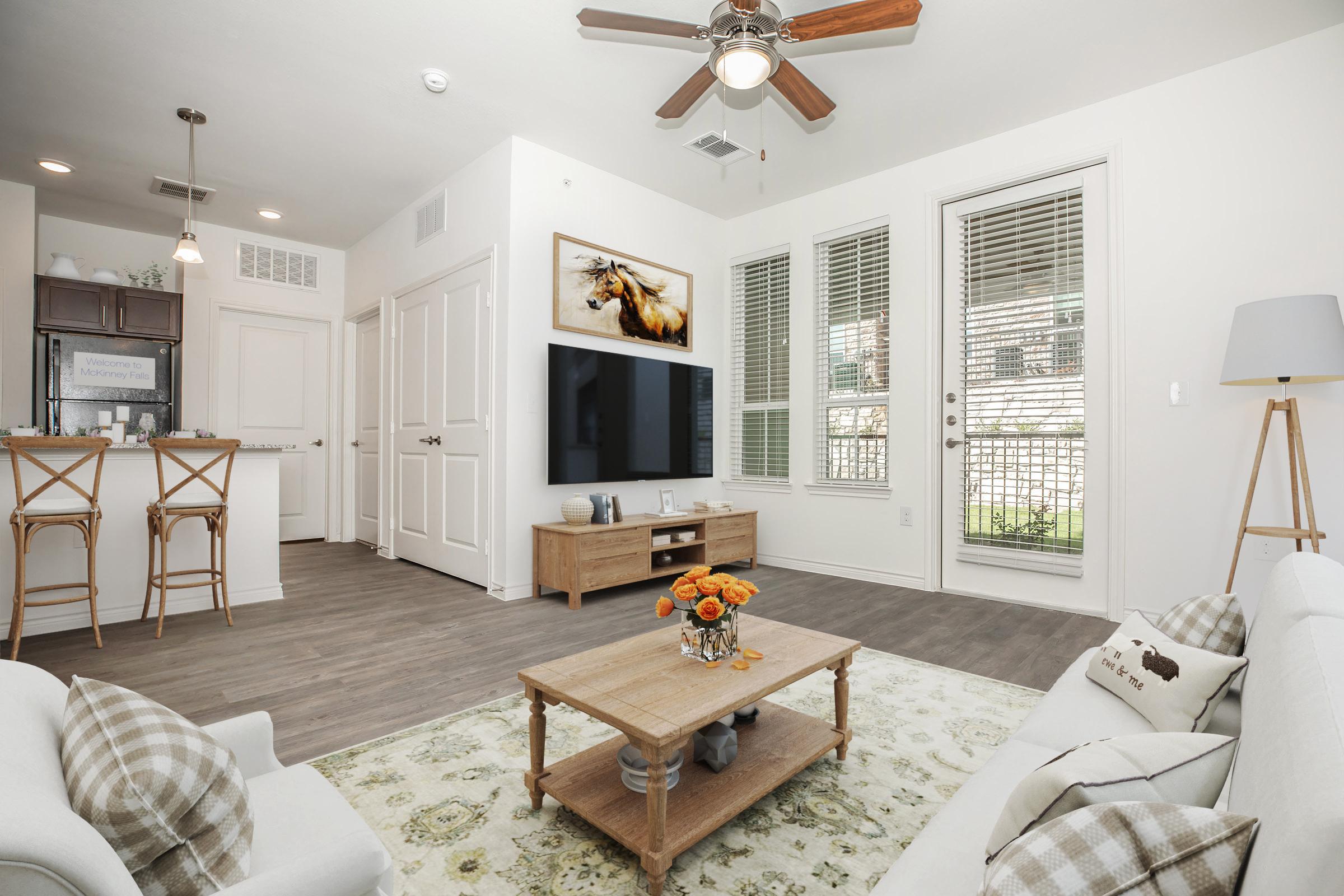 A modern living room featuring a light-colored sofa with checkered pillows, a wooden coffee table with a vase of flowers, and a large flat-screen TV mounted on the wall. There is a dining area with bar stools and a kitchen in the background. Sunlight streams through large windows with blinds.
