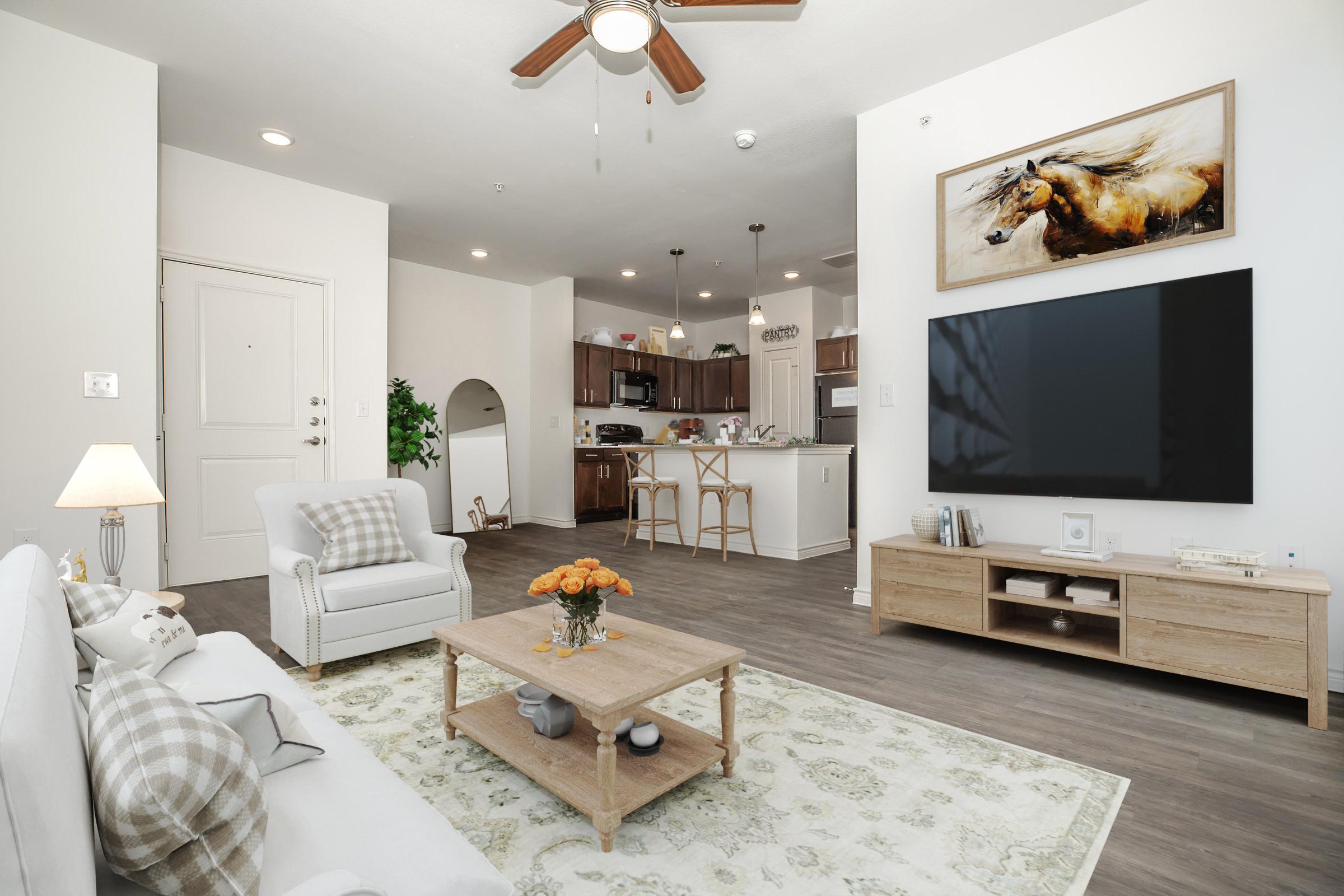A cozy living room featuring a white sofa with decorative pillows, a wooden coffee table with a vase of flowers, and a large flat-screen TV mounted on the wall. The space includes a small dining area with bar stools, and the kitchen can be seen in the background. Bright and inviting, with a modern aesthetic.