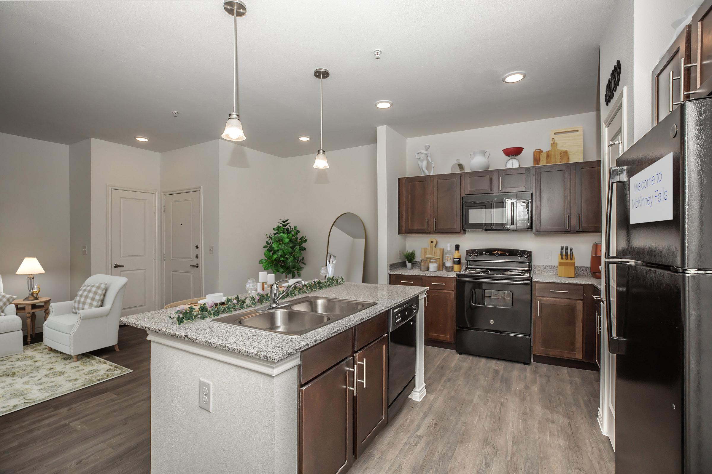 Modern kitchen with dark wood cabinets, stainless steel appliances, and a granite countertop. Features a central island with a sink, pendant lighting, and decorative plants. In the background, there is a comfortably furnished living area with a white chair and a rug, along with a door leading to the hallway.