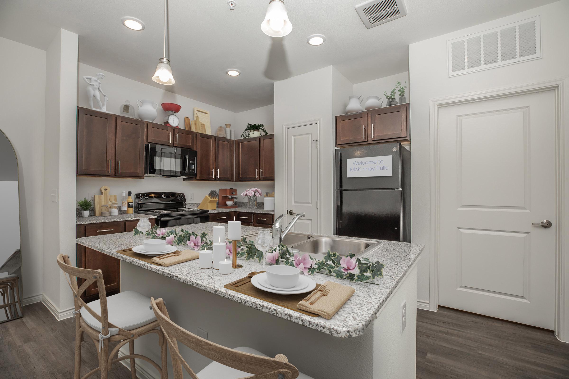 A modern kitchen featuring dark brown cabinetry, a stainless steel refrigerator, and a microwave. The island includes a countertop with decorative plates, napkins, and faux flowers. Light fixtures hang from the ceiling, and the space is bright with various decorative items on the shelves. A welcoming sign is visible on the wall.