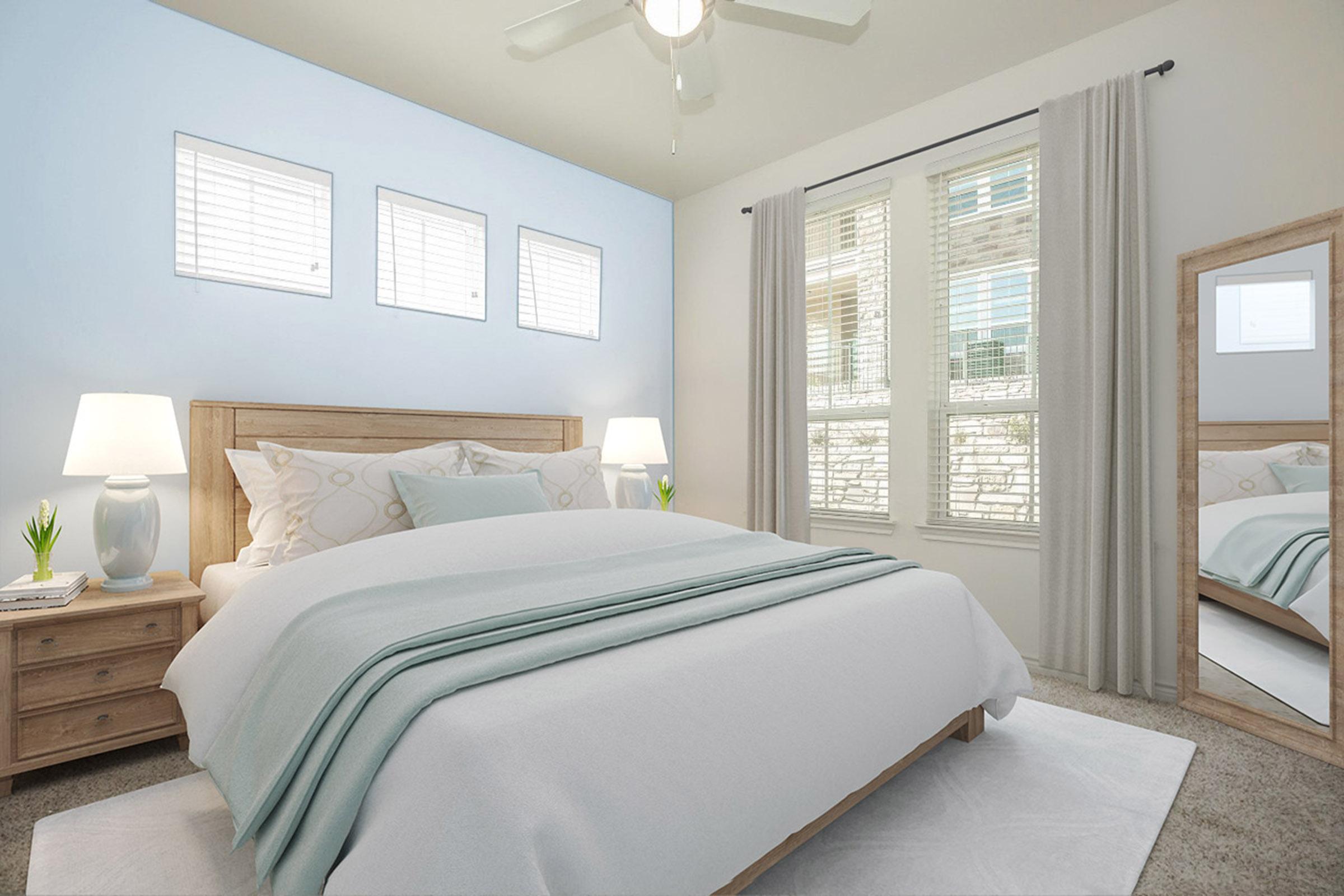 A bright and airy bedroom featuring a light wood bed frame with white bedding, two bedside tables with lamps, and a potted plant on one table. A large mirror stands next to a window with white curtains. The walls are painted light blue, and there are three small windows letting in natural light.